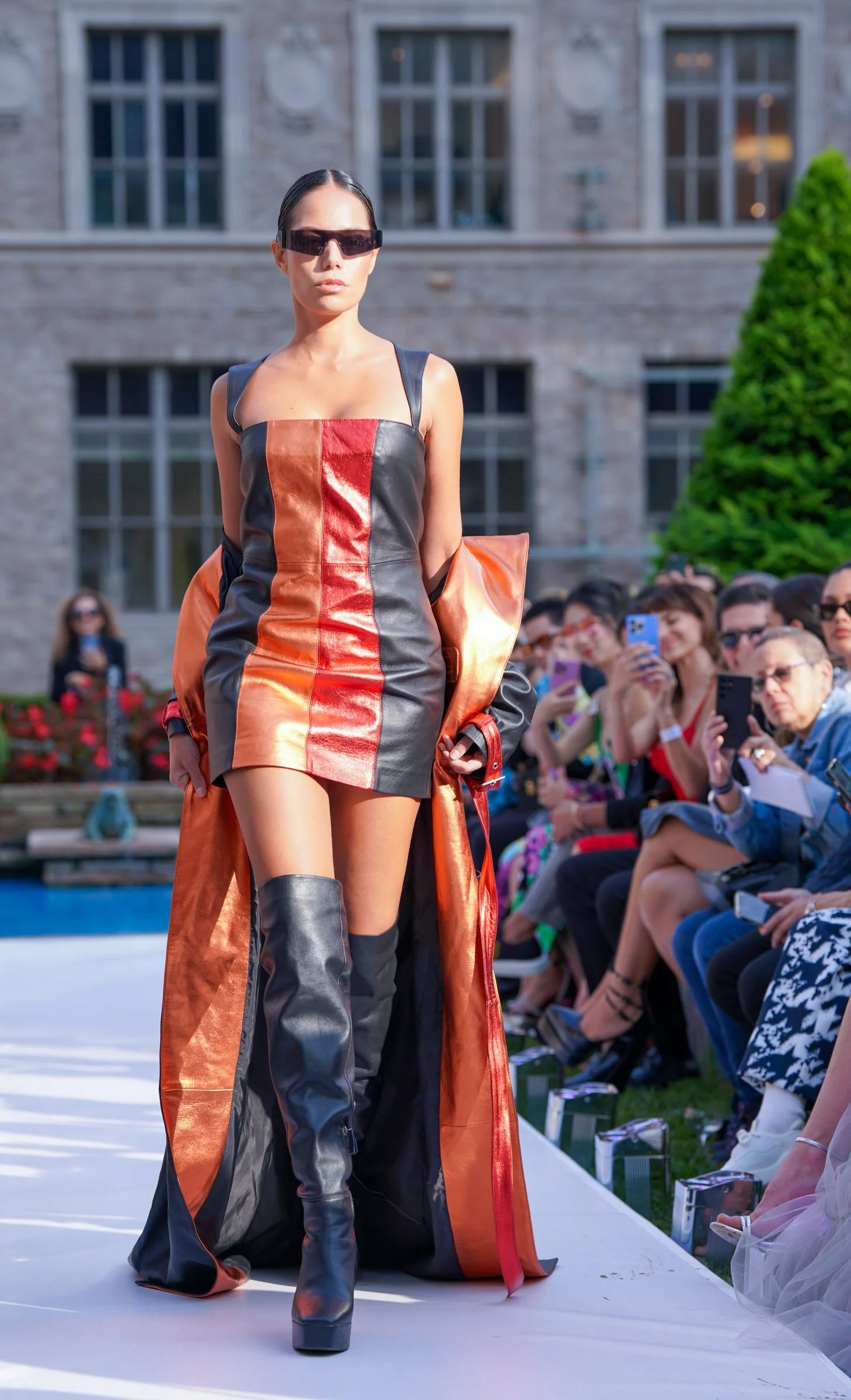 A female model walking on an outdoor runway at a fashion show, wearing a black, orange, and red leather dress with thigh-high black boots and oversized sunglasses, with an audience seated on the side taking photos.