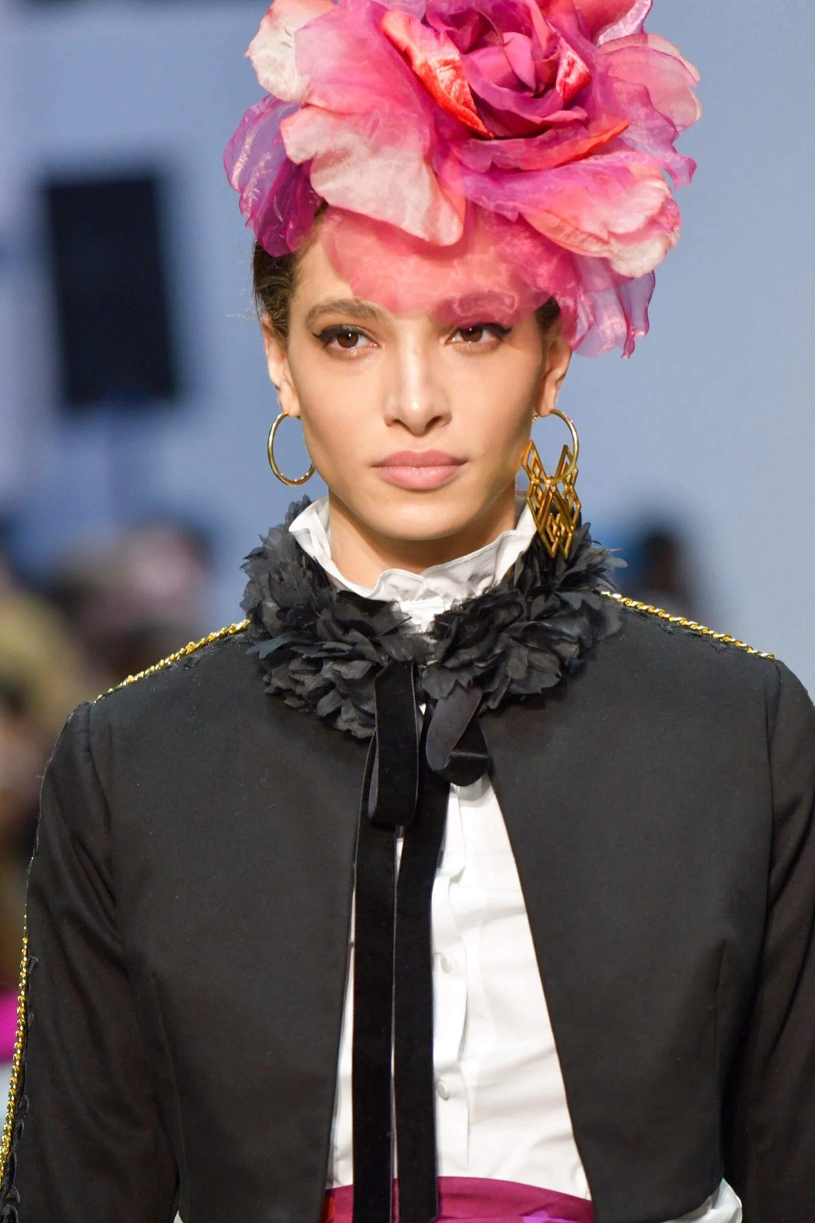 A woman wearing a large pink and purple flower headpiece, black suit jacket with gold details, and hoop earrings, standing indoors against a blue background after a flamenco dance.