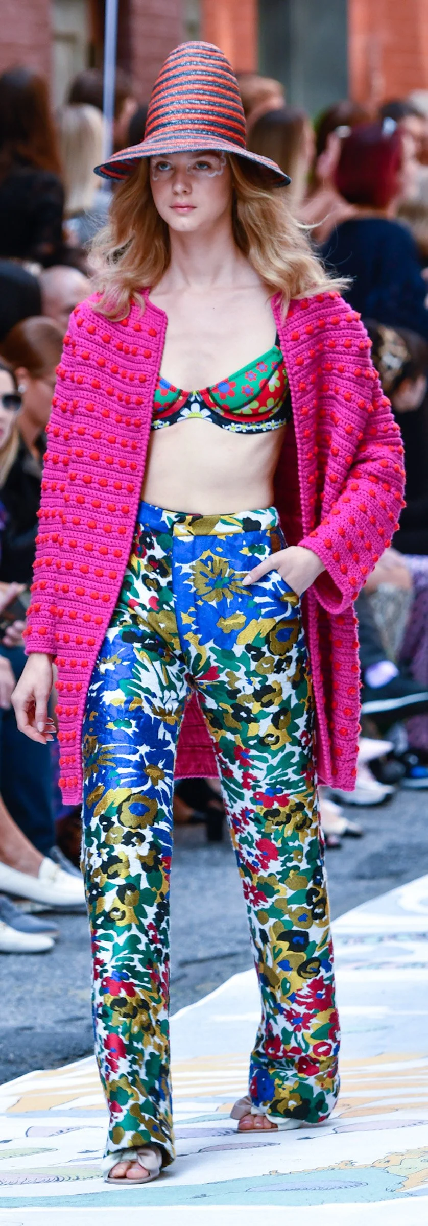 Fashion model walking on runway in colorful, patterned clothing including a large striped hat, pink textured coat, floral bra, and wide-leg floral pants, with audience in background. Designer, Cynthia Rowley.