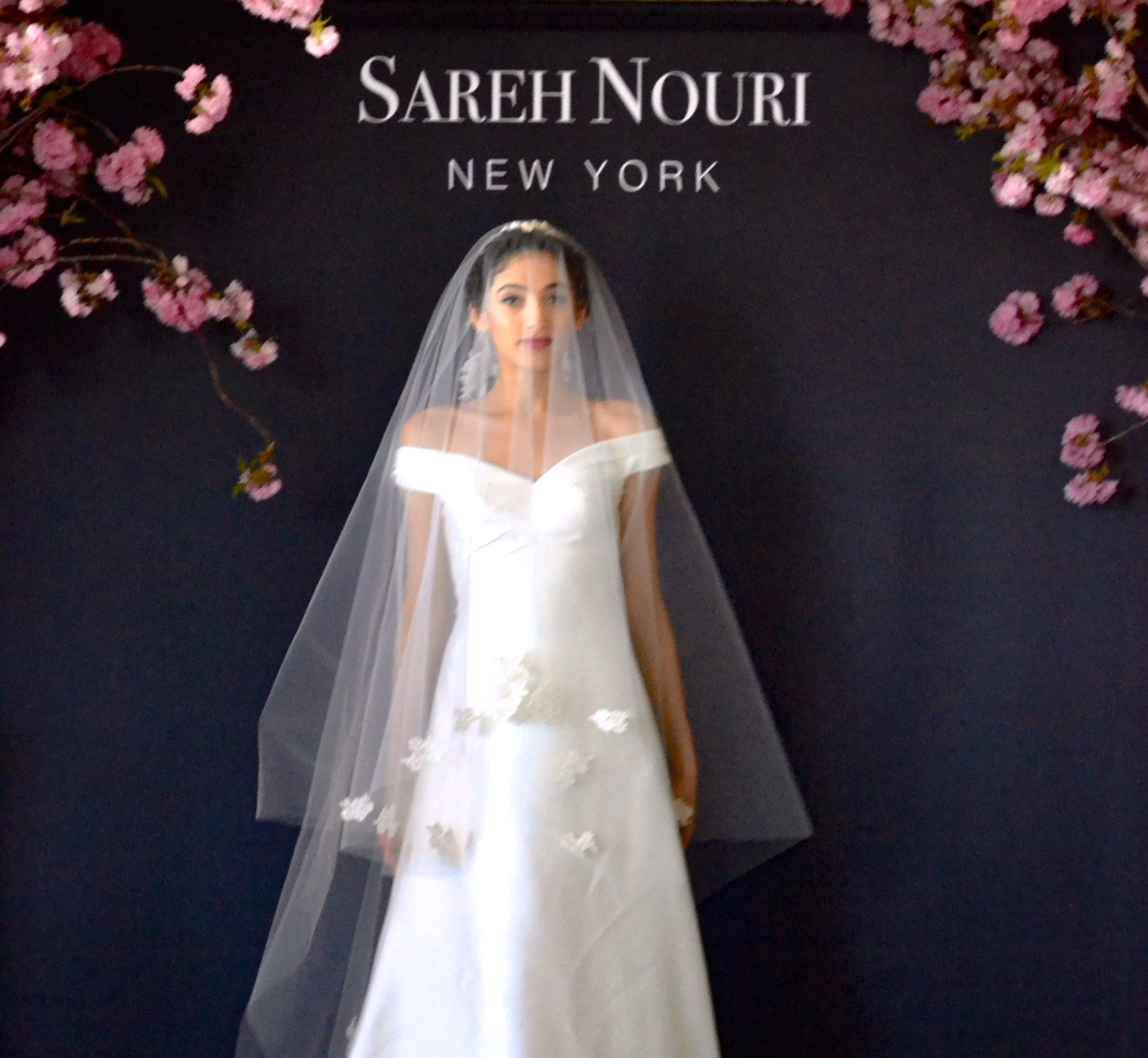 A woman dressed in a white gown and veil, standing in front of a black backdrop with pink flowers on either side. The backdrop has the words 'SAREH NOURI NEW YORK' written on it.
