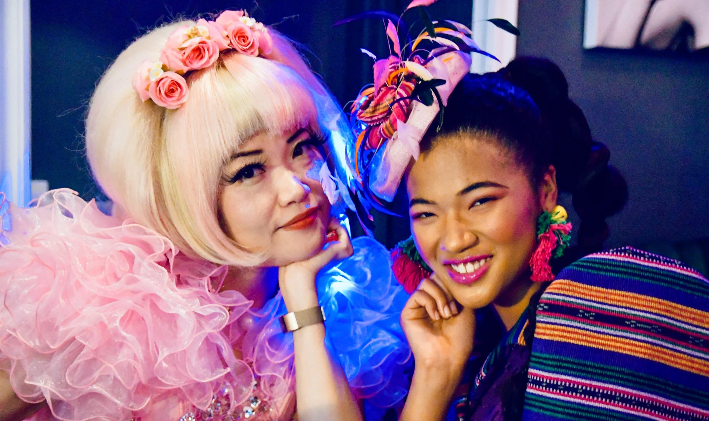 Two women smiling, one with pink hair, wearing a pink dress with ruffled details and a pink rose headband, the other with dark hair in a bun, wearing a striped colorful sweater and fringed earrings, close together with their hands near their faces.