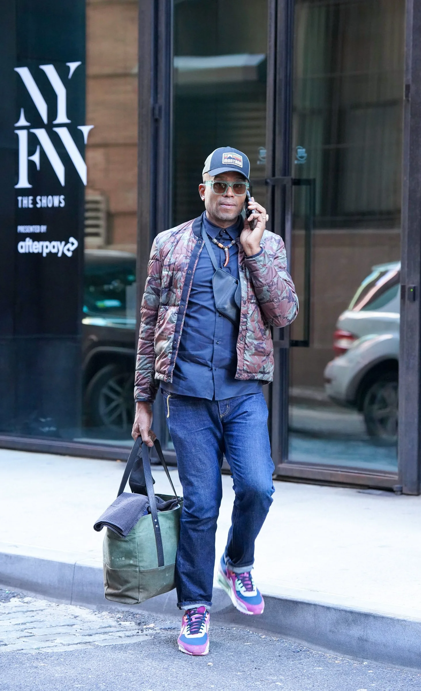 Man walking on sidewalk, talking on cell phone, carrying a duffel bag, wearing sunglasses, baseball cap, jeans, colorful sneakers, and a patterned jacket, outside a glass building.