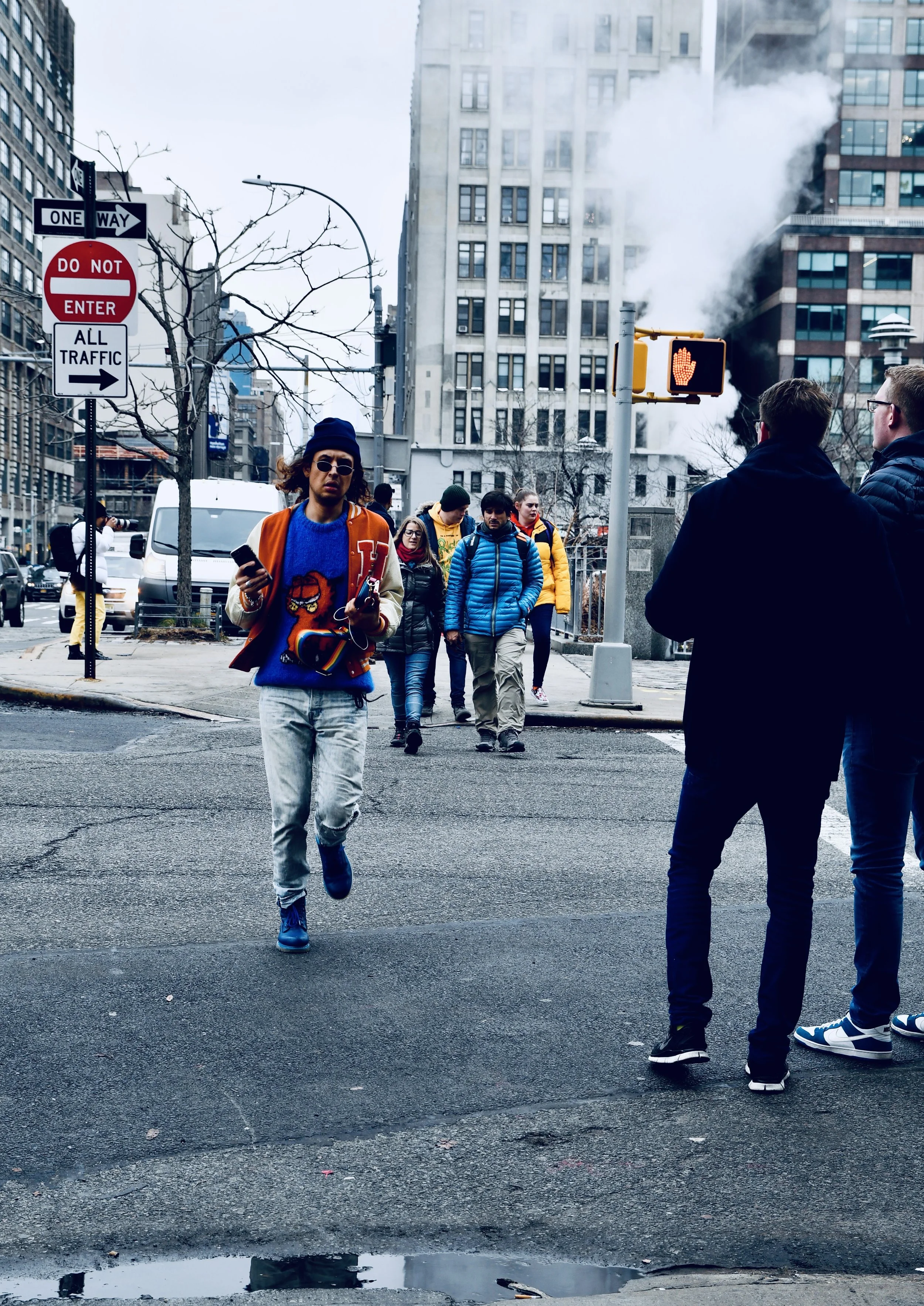 Pedestrians crossing streets in a busy city intersection with a tall building emitting steam or smoke in the background and traffic signals visible.