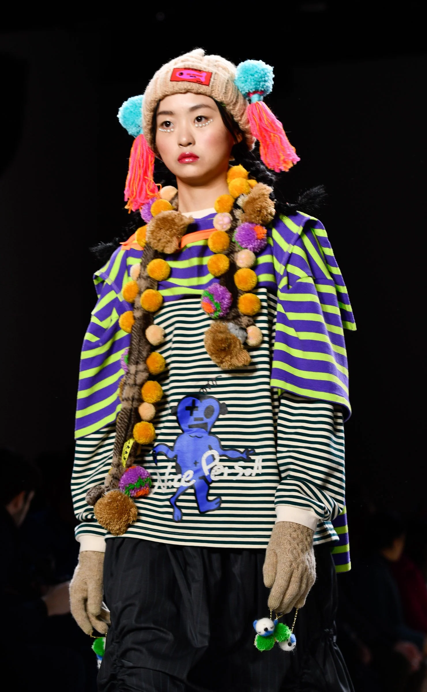 A fashion model wearing a colorful, layered outfit with striped clothing, a bear-themed knitted hat with pink tassels, and expressive accessories including pom-pom necklaces and plush toys, walking on a runway with a black background.