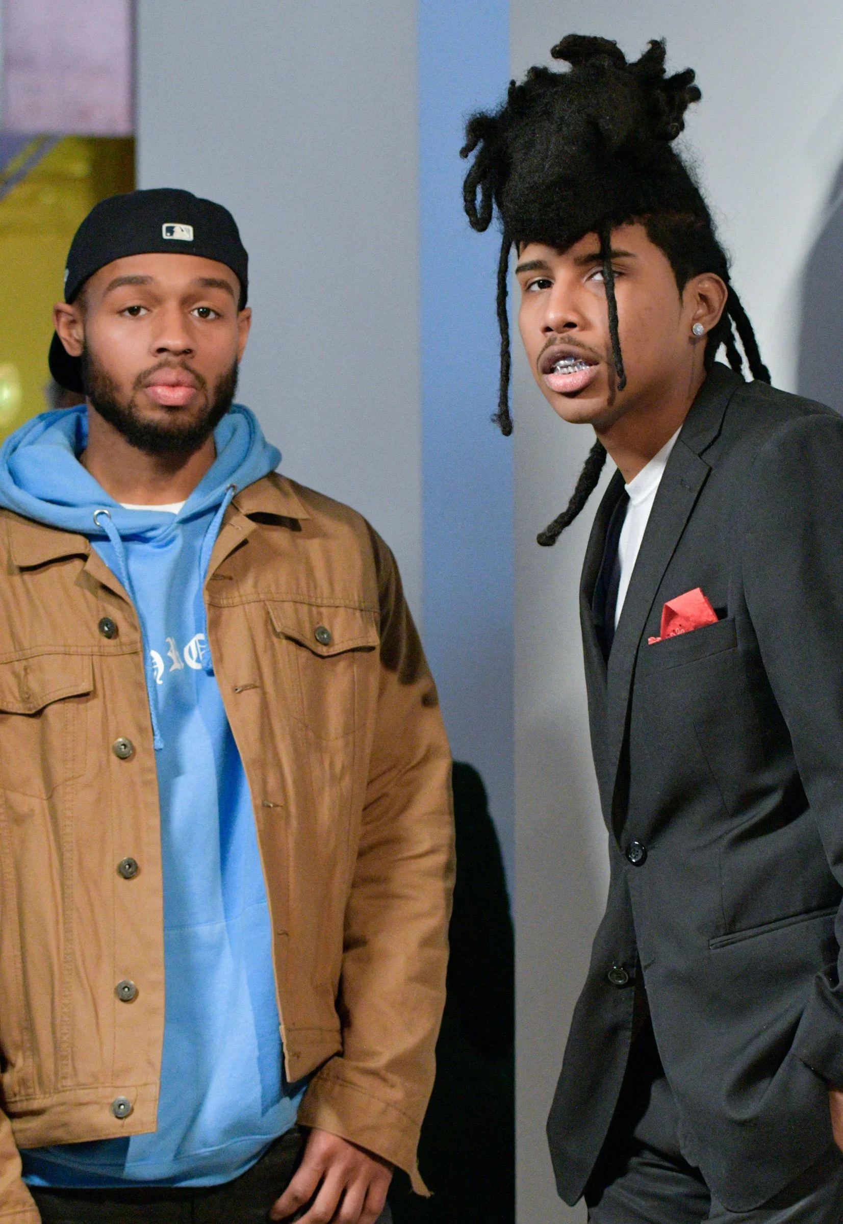 Two men standing side by side in an indoor setting, one wearing a baseball cap backwards, a brown jacket, and a blue hoodie, the other wearing a black suit with a white shirt, earrings, and sporting dreadlocks.