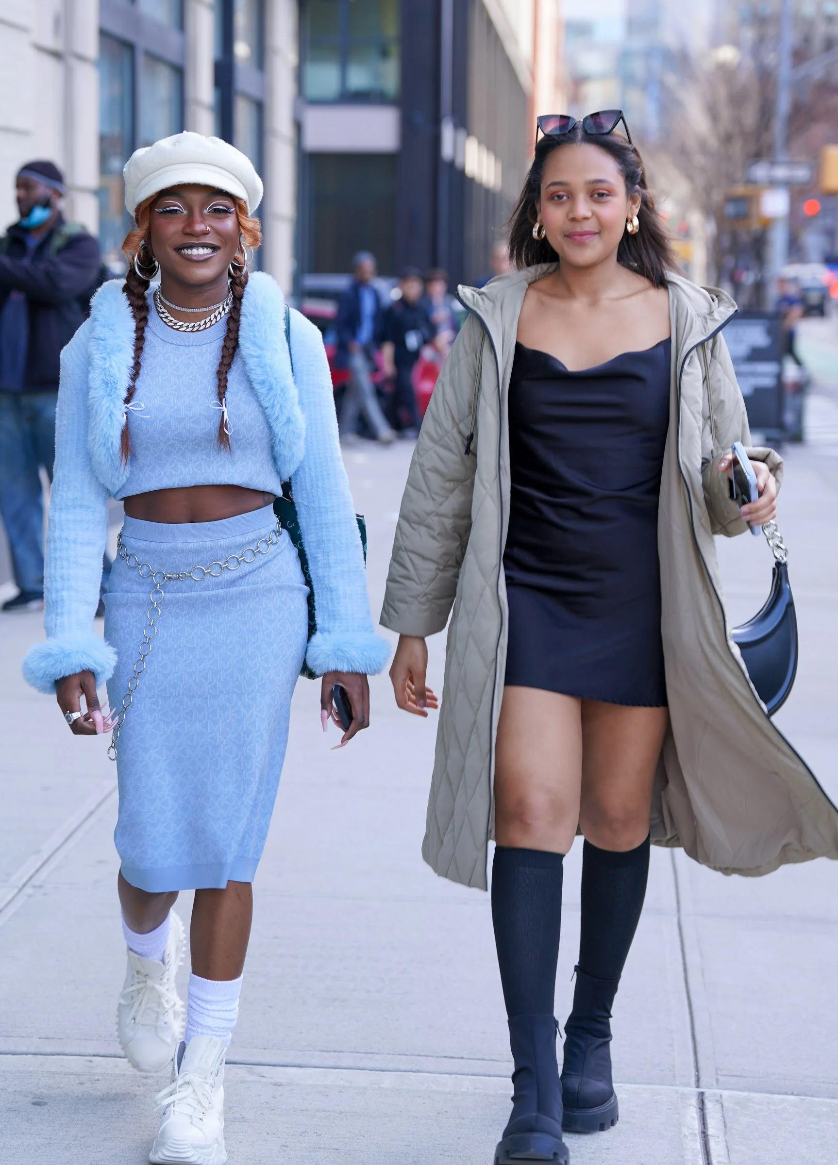 Two young women walking on a city sidewalk, smiling, dressed in trendy fashion. One wears a white beret and light blue outfit with chunky sneakers, and the other wears a beige jacket over a black dress with knee-high socks and chunky black boots.
