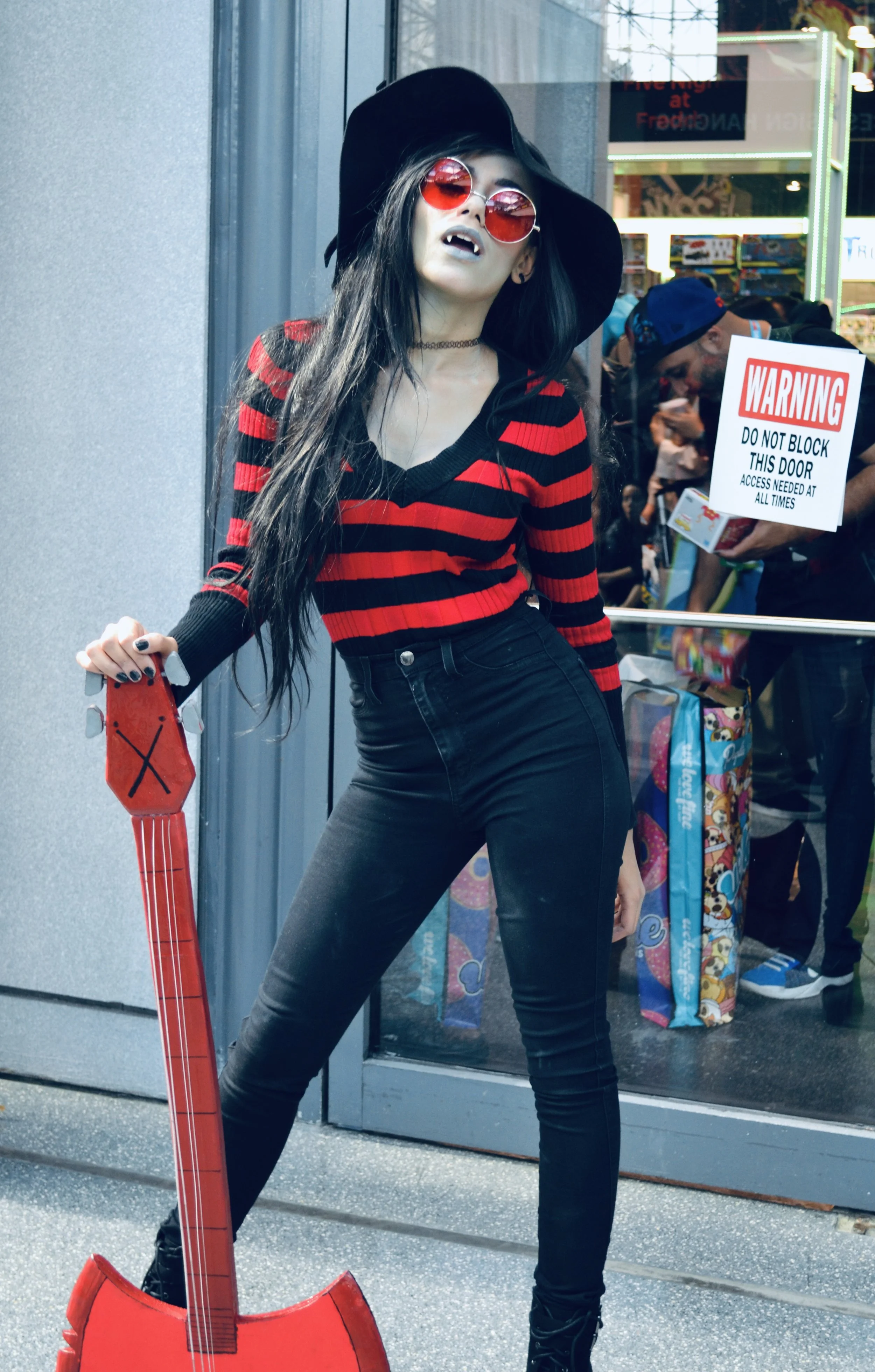 A young woman with long black hair wearing red-tinted round sunglasses, a wide-brimmed black hat, a red and black striped sweater, black jeans, and black boots, posing outdoors with a red inflatable guitar in front of a glass storefront.