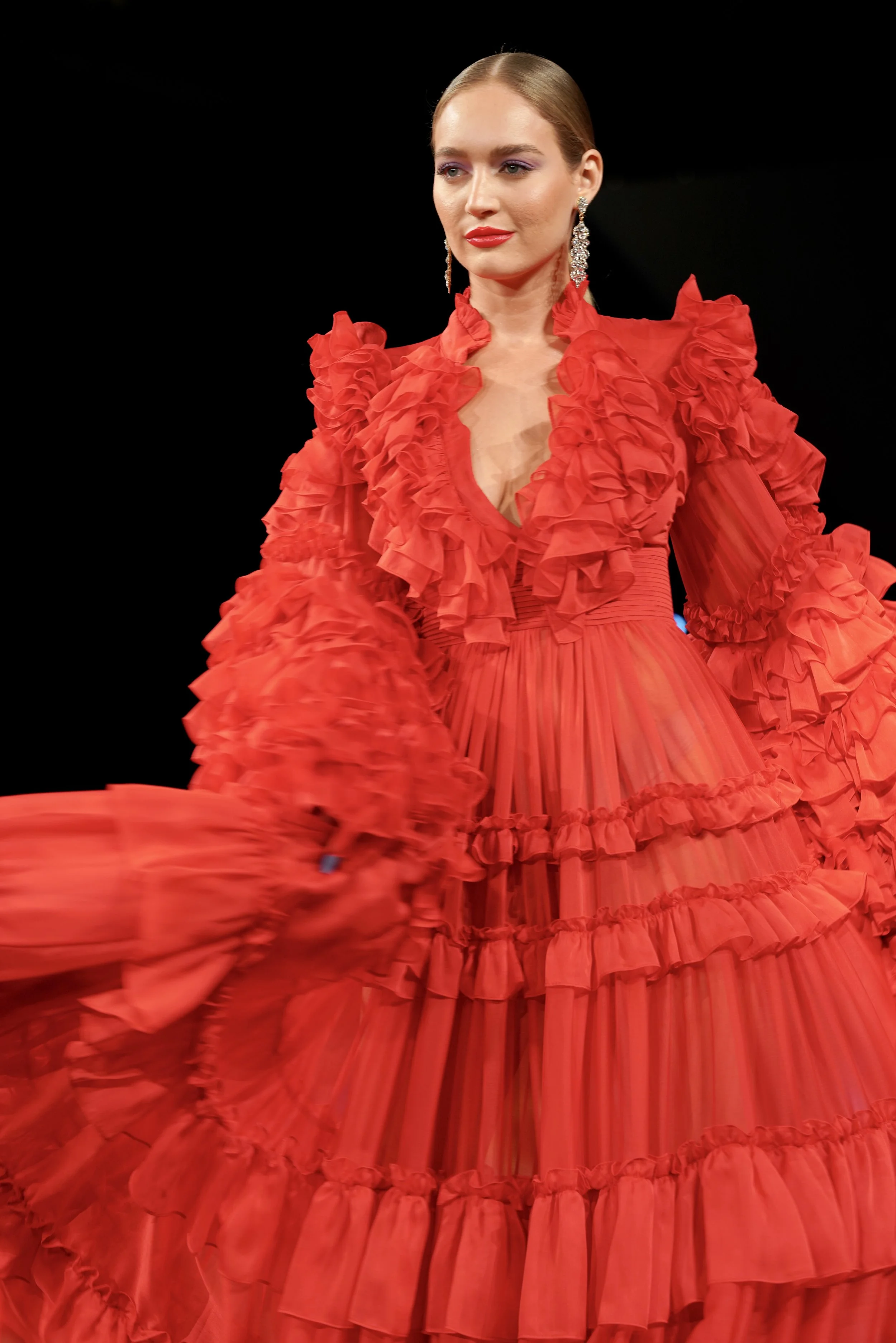 Woman in a red dress with ruffled details, long pleated skirt, and large earrings, against a black background.