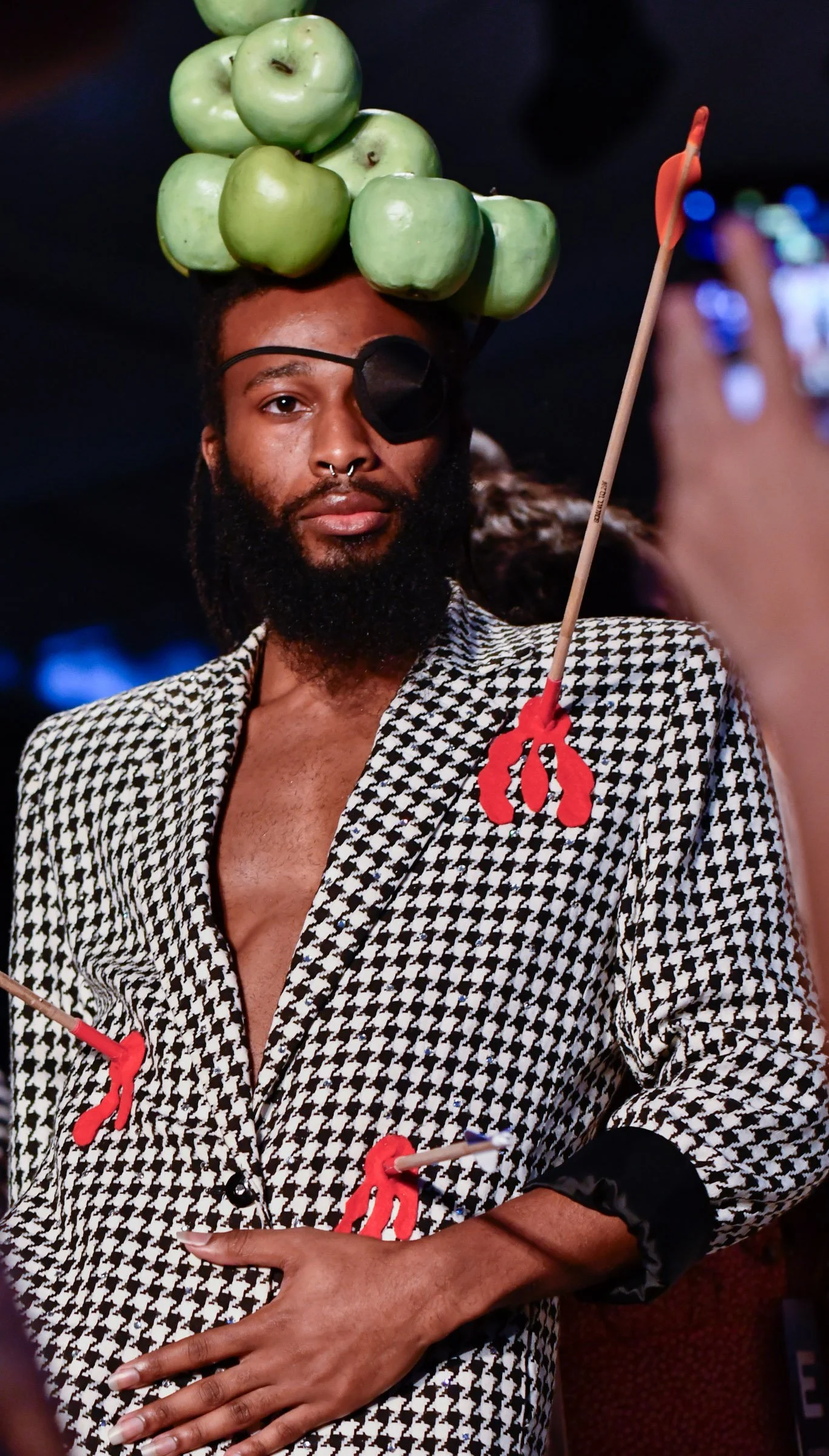 A man with a beard, wearing an eye patch over his right eye and a black eyepatch on his forehead, takes a selfie. He is dressed in a black and white houndstooth patterned blazer with a deep neckline. His head is topped with a large cluster of green a