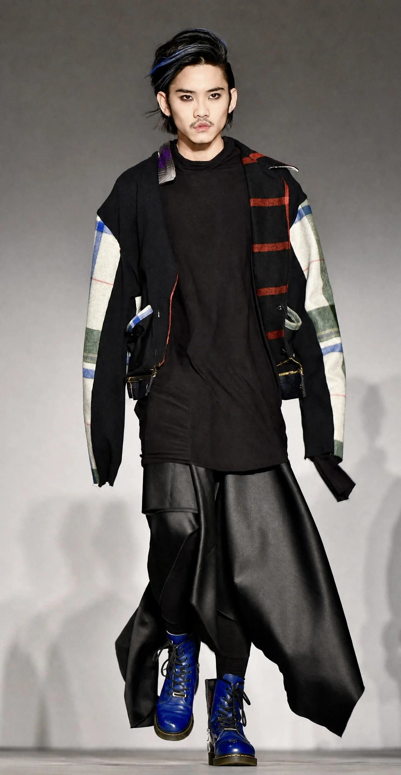 A male model walking down a runway wearing a layered black top, a colorful patchwork jacket, black wide-leg leather pants, and bright blue combat boots.