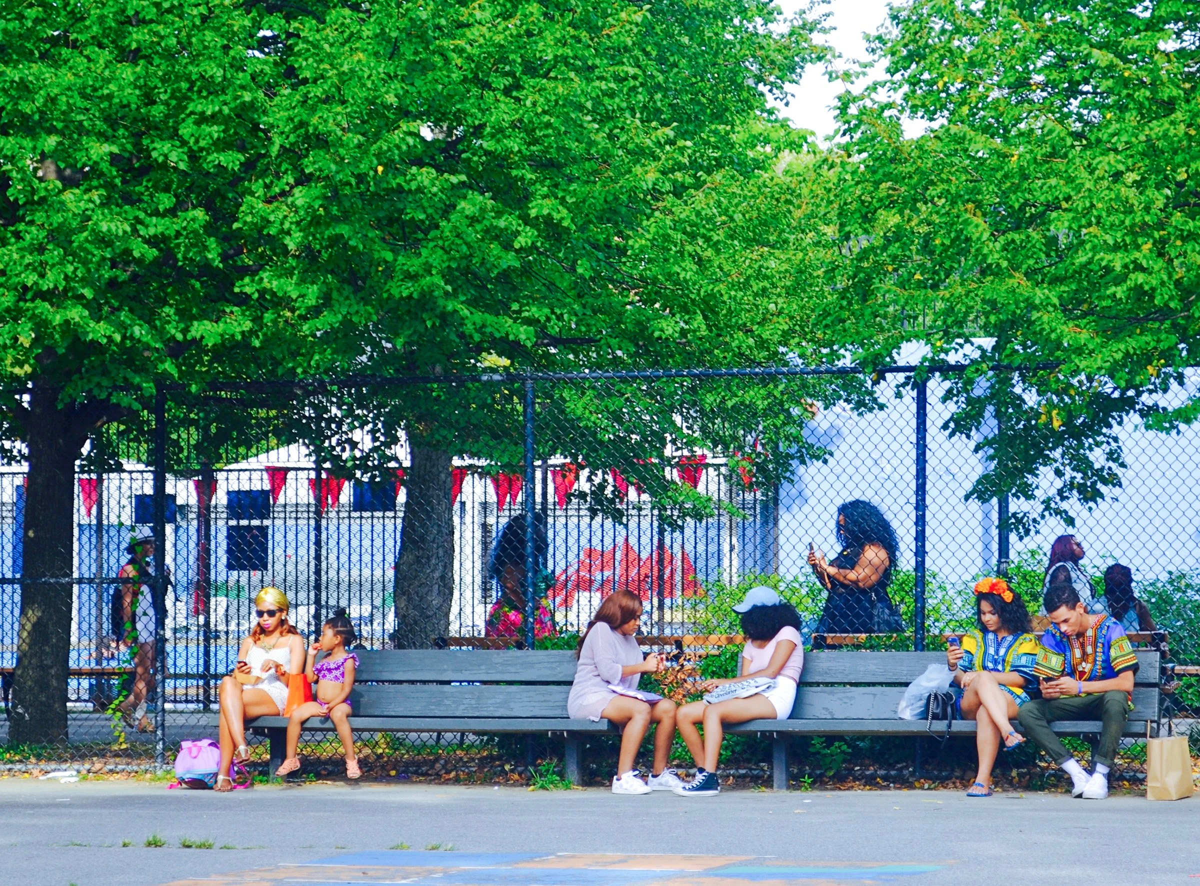 People sitting on a bench and standing near a chain-link fence with trees behind them at an outdoor event.