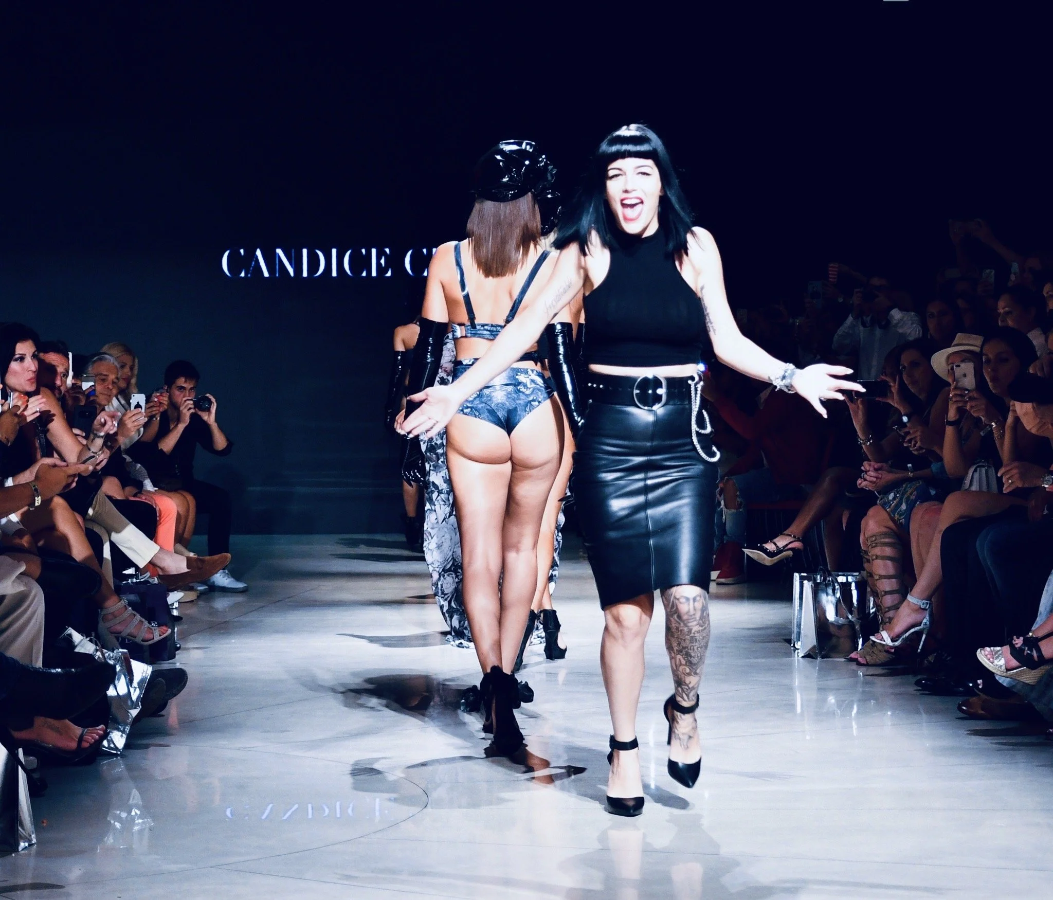 Models walking down runway at fashion show with audience seated on both sides, some taking photos, and a sign displaying 'CANDICE CUOCO'