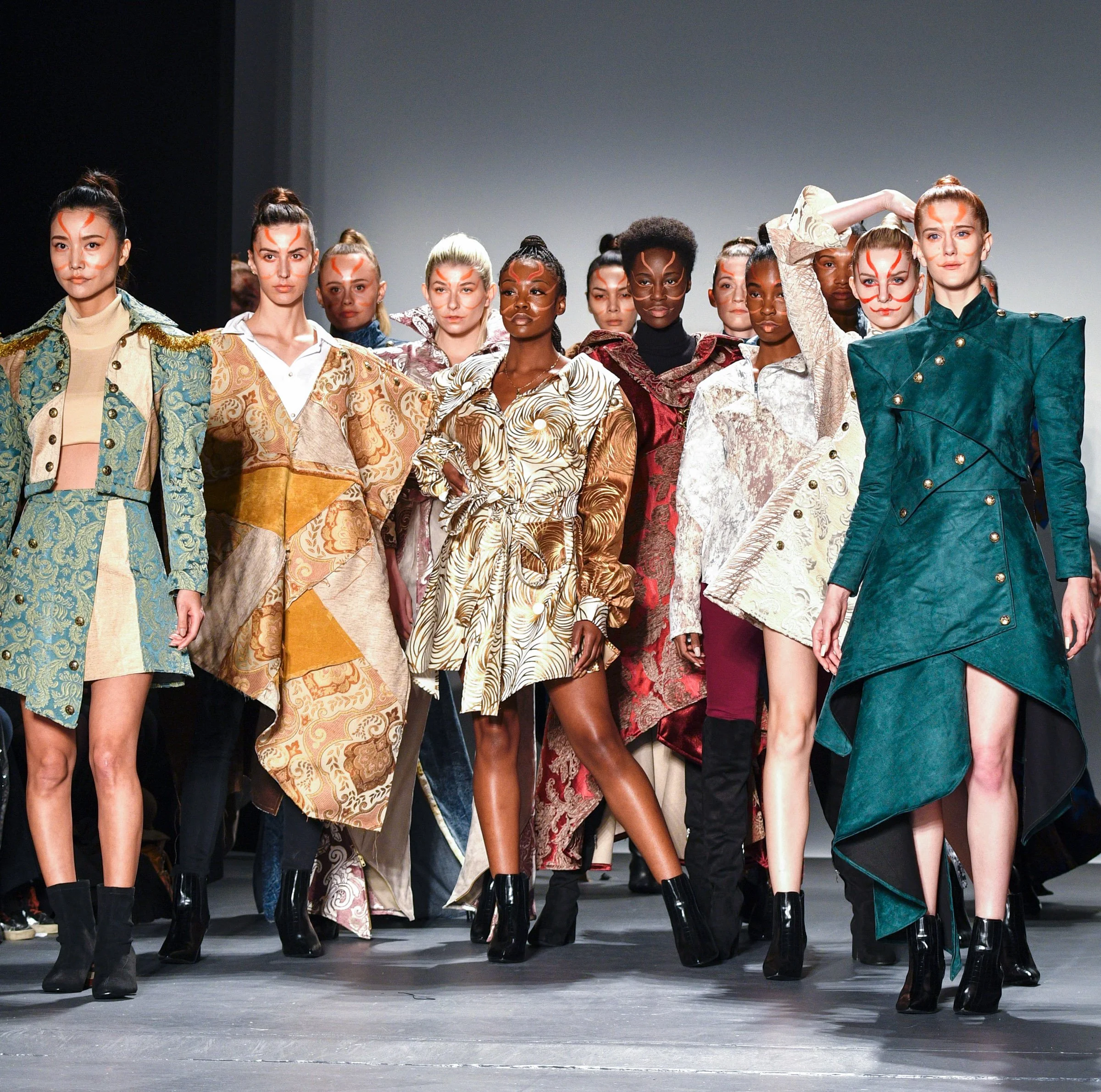Models walking on a runway during a fashion show, wearing designer outfits with bold makeup and accessories.