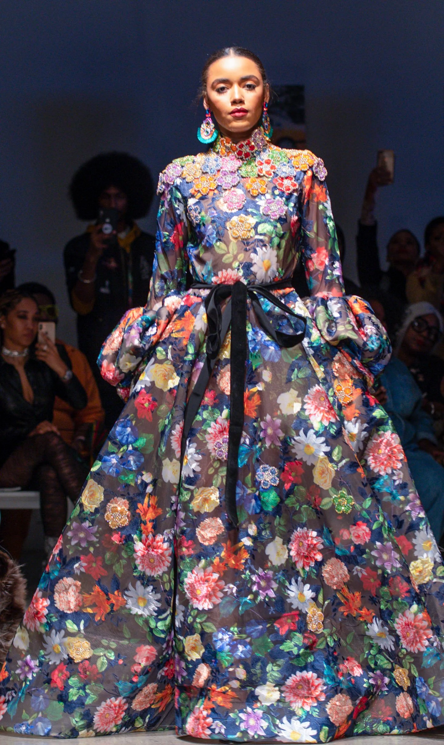A model walking down the runway in a colorful floral gown with a black ribbon at the waist, accessorized with matching large floral earrings.
