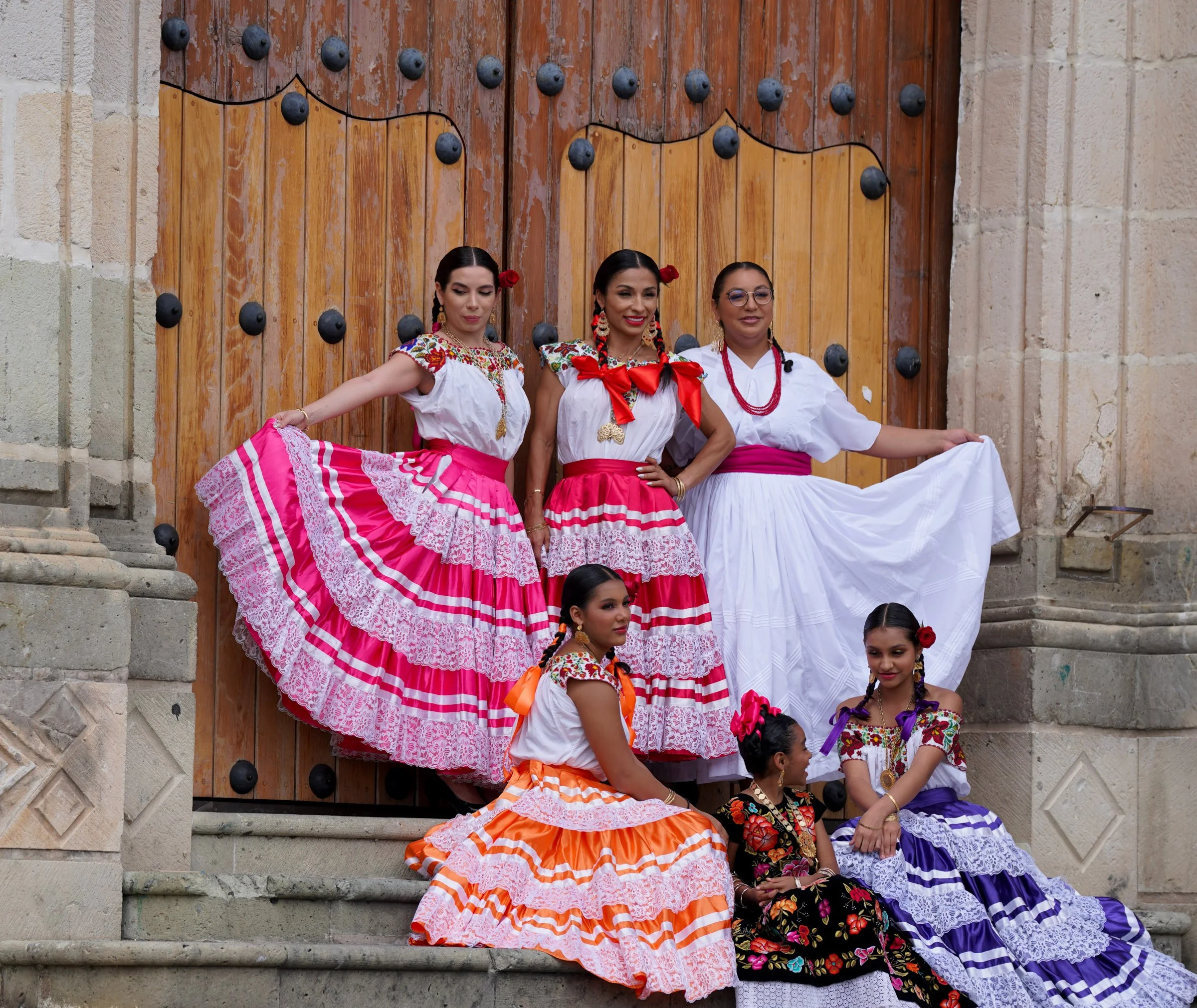 Group of women in traditional Mexican dresses performing a dance on the steps of an old stone building with a large wooden door in the background. Oaxaca, Mexico.