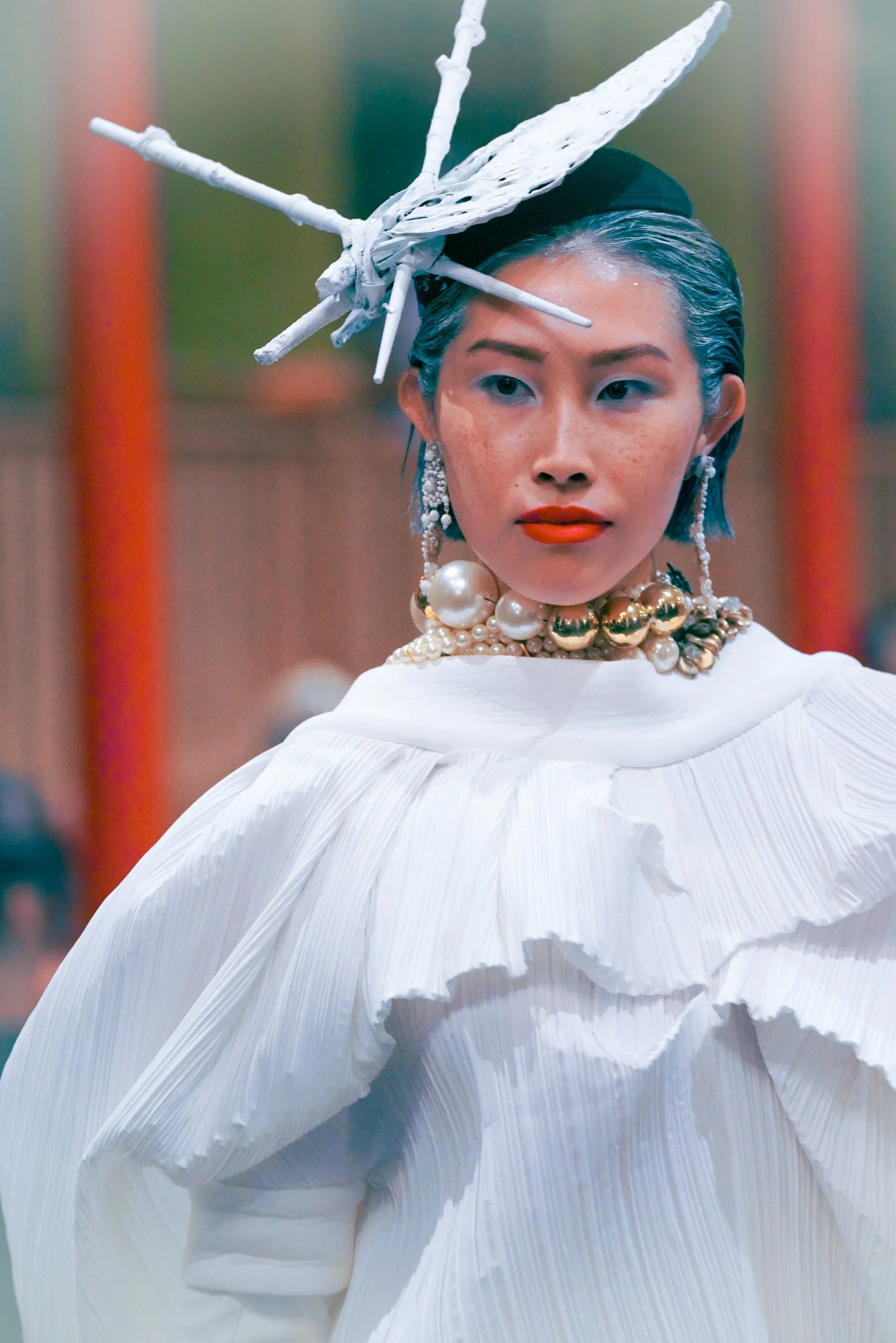A woman with gray hair styled back, wearing large pearl and gold jewelry, an elaborate white pleated dress, and a headpiece resembling a white insect or butterfly, in a fashion-forward or artistic pose.