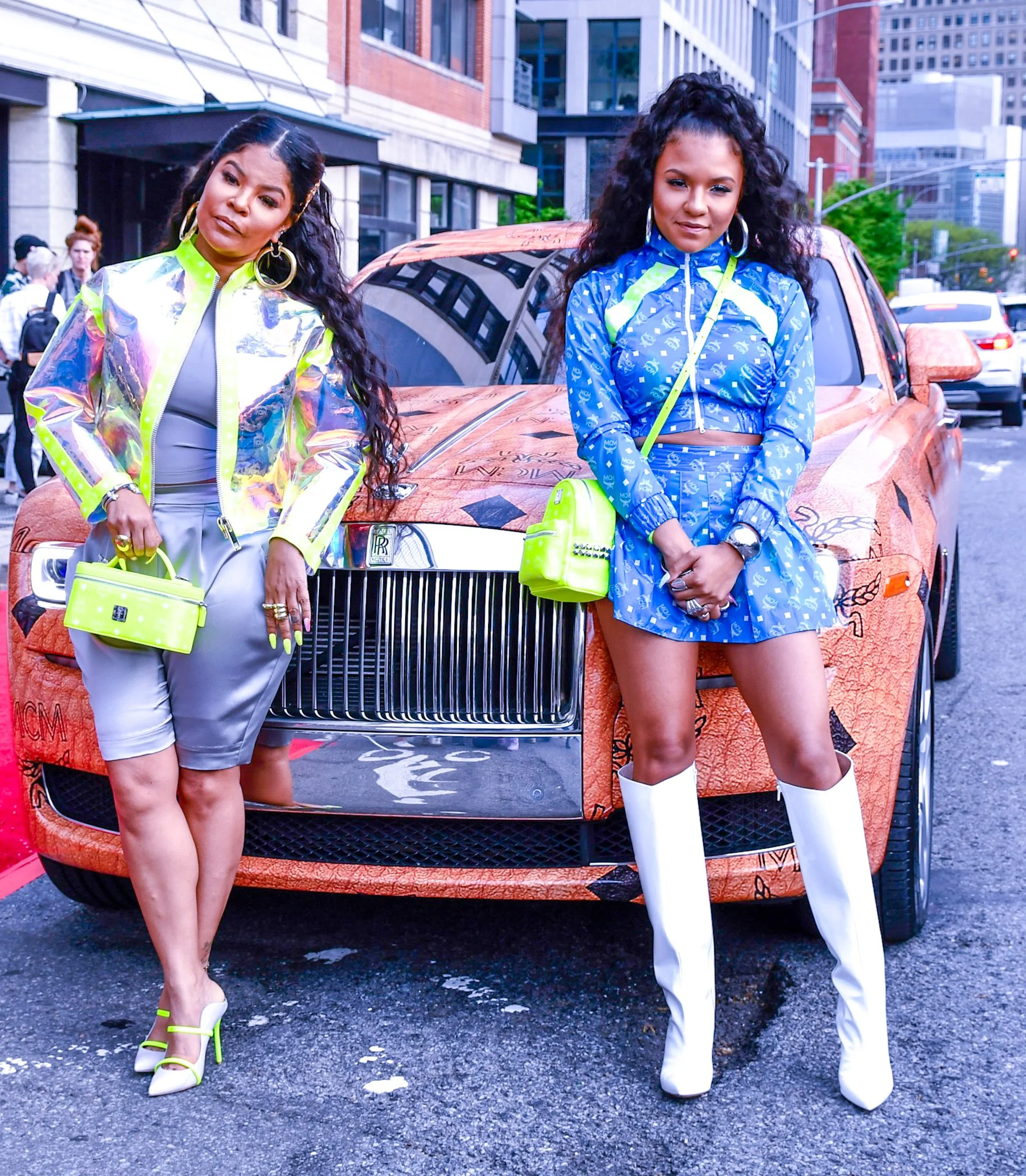 Two women standing in front of a luxury car on a city street, wearing brightly colored outfits and high heels.