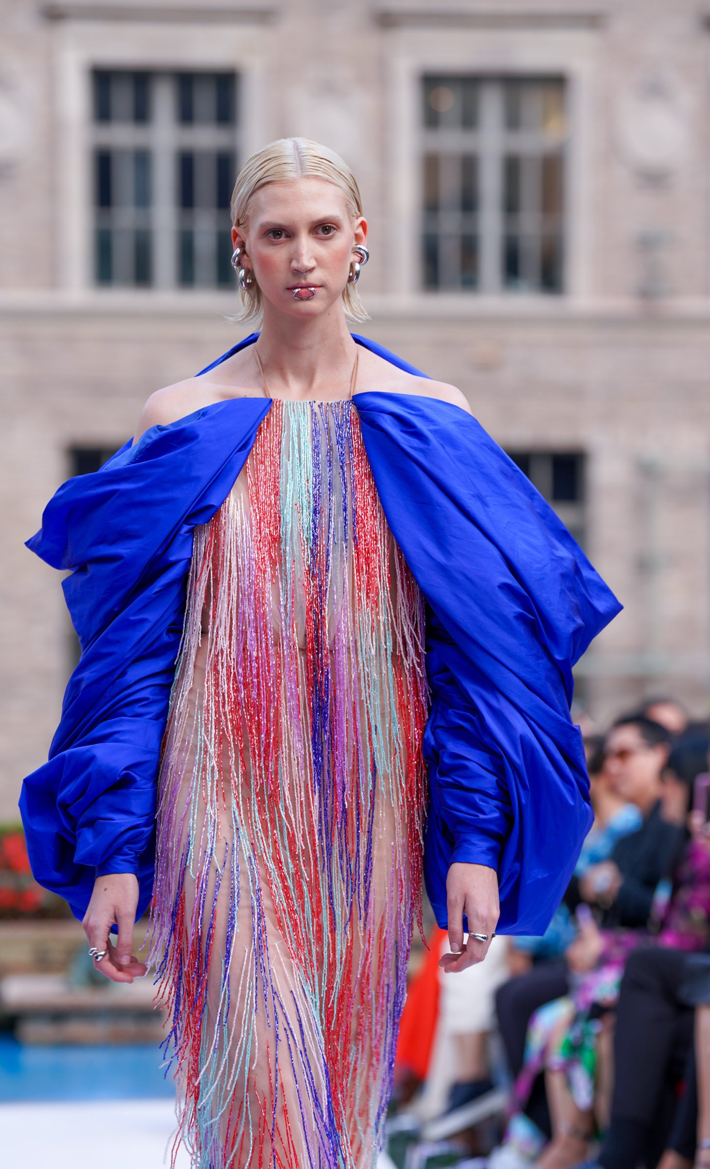 A fashion model walking down an outdoor runway, wearing a colorful dress with vertical streamers in shades of red, pink, orange, purple, and blue, topped with a bright blue oversized jacket.