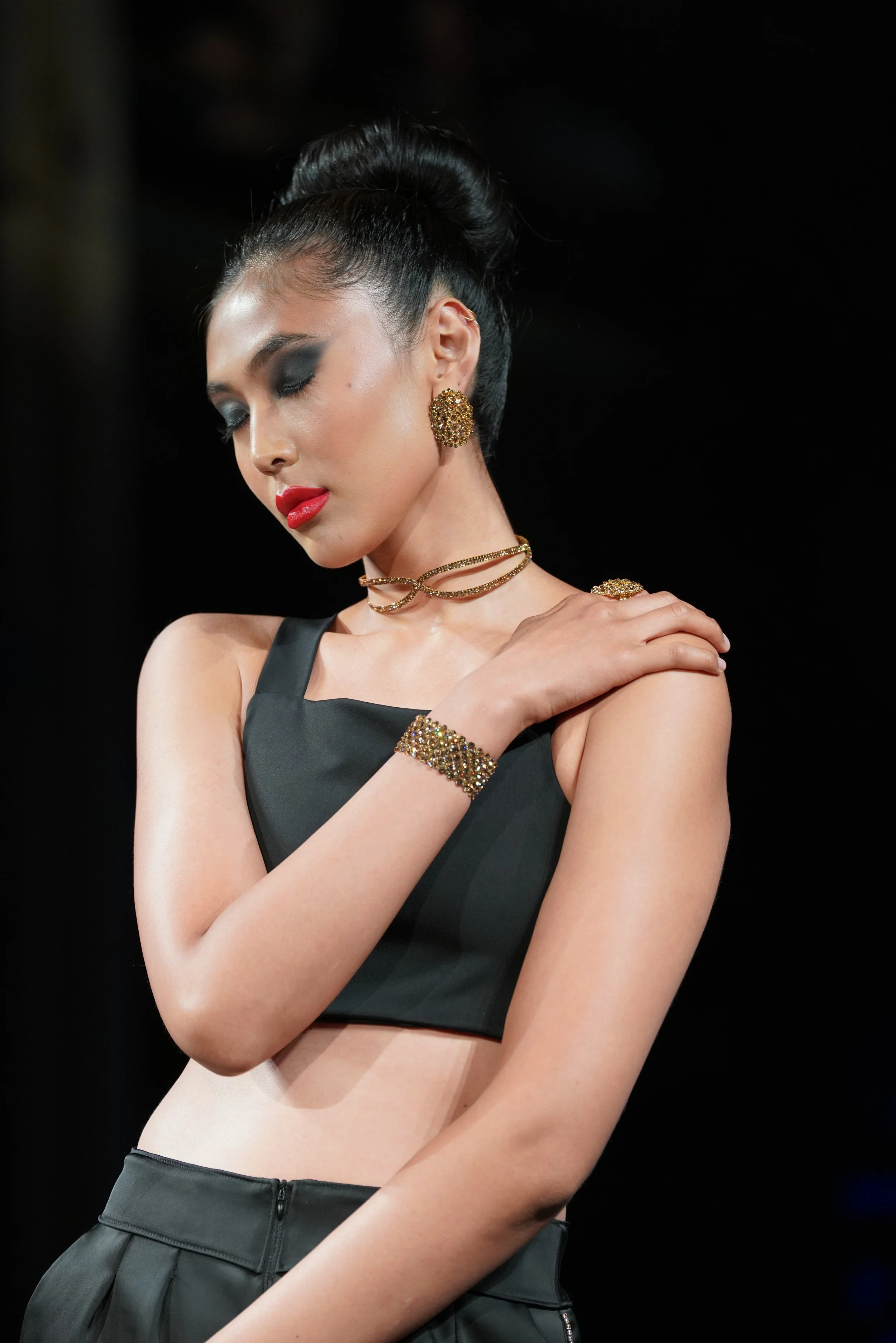 A woman with dark hair styled in an elegant updo, wearing black eye makeup, red lipstick, gold jewelry including earrings, choker, ring, bracelet, and shoulder embellishment, dressed in a black sleeveless top and black high-waisted skirt, standing wi