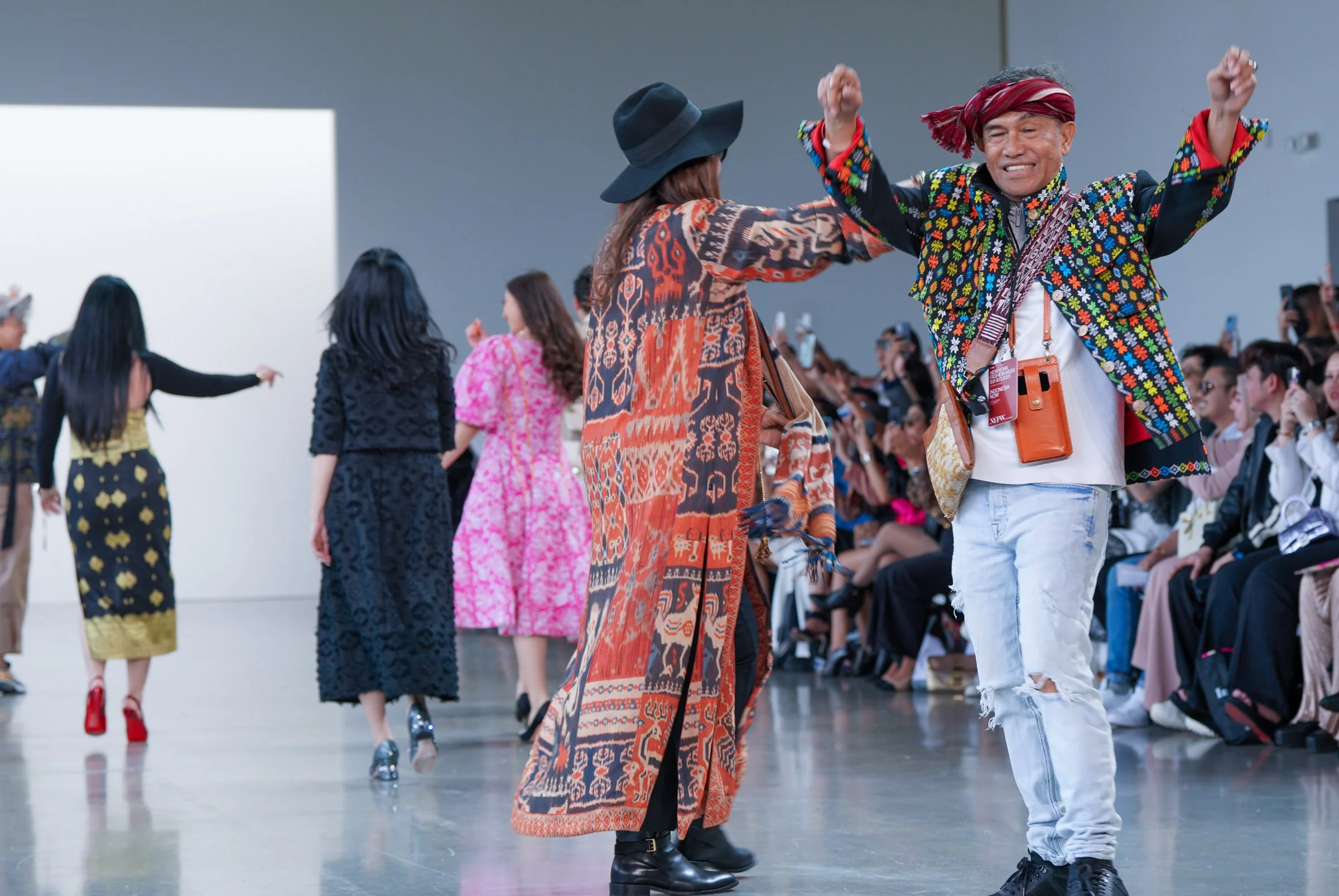 People walking on a runway during a fashion show, wearing colorful and patterned outfits, with an audience capturing the moment on their phones.