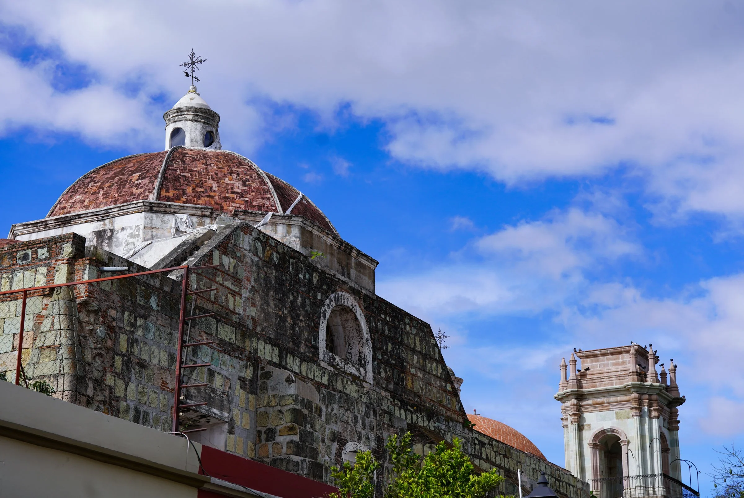 A historic church with a stone facade and a red-tiled dome, topped with a weather vane, against a bright blue sky with scattered clouds. Oaxaca, Mexico.