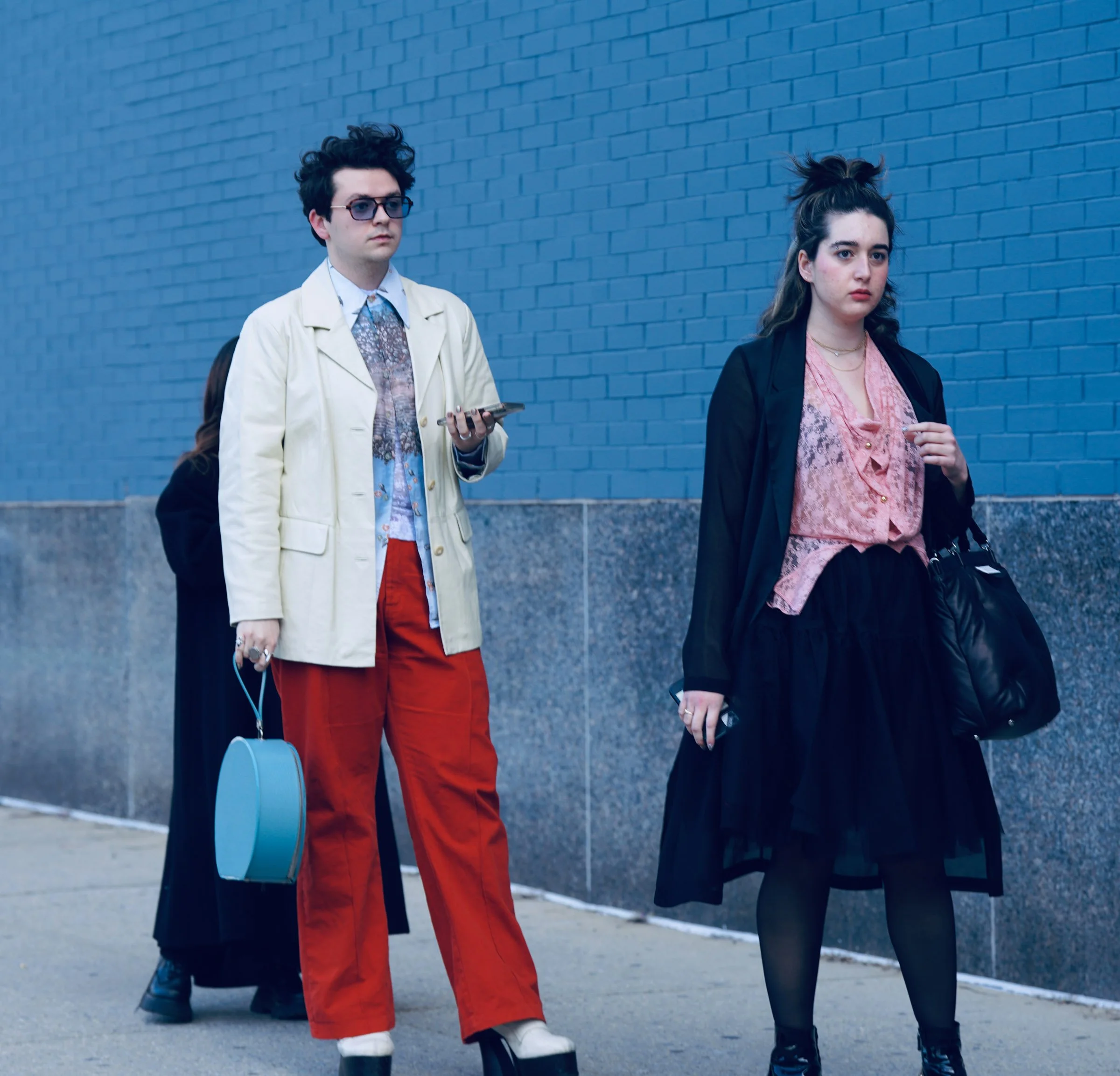 Two young adults walking on a sidewalk against a blue brick wall. The person on the left wears glasses, a light-colored jacket, a patterned shirt, red pants, and white shoes. They are holding a phone and have earphones plugged into their ears. The pe