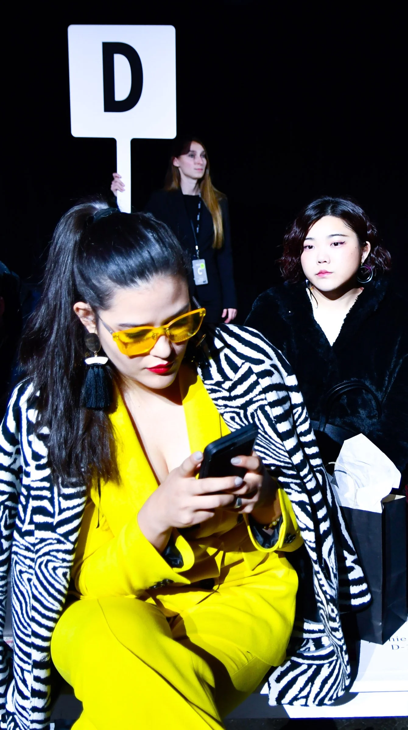 A woman in a bright yellow suit with zebra-patterned accents is sitting and looking at her phone. Behind her, a woman holding a sign with a large letter D is standing with others seated nearby in a dark environment. Front row socialites.