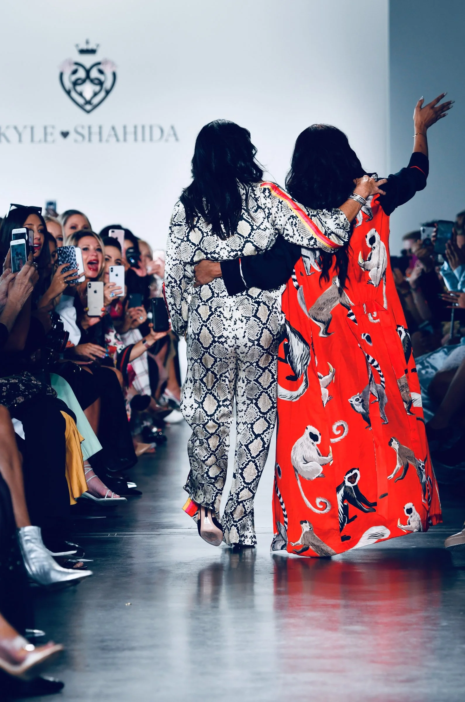 Fashion models walking the runway at a fashion show, with an audience taking pictures. The background features a logo with a crown and text that reads 'SKYLE SHAHIDA'.