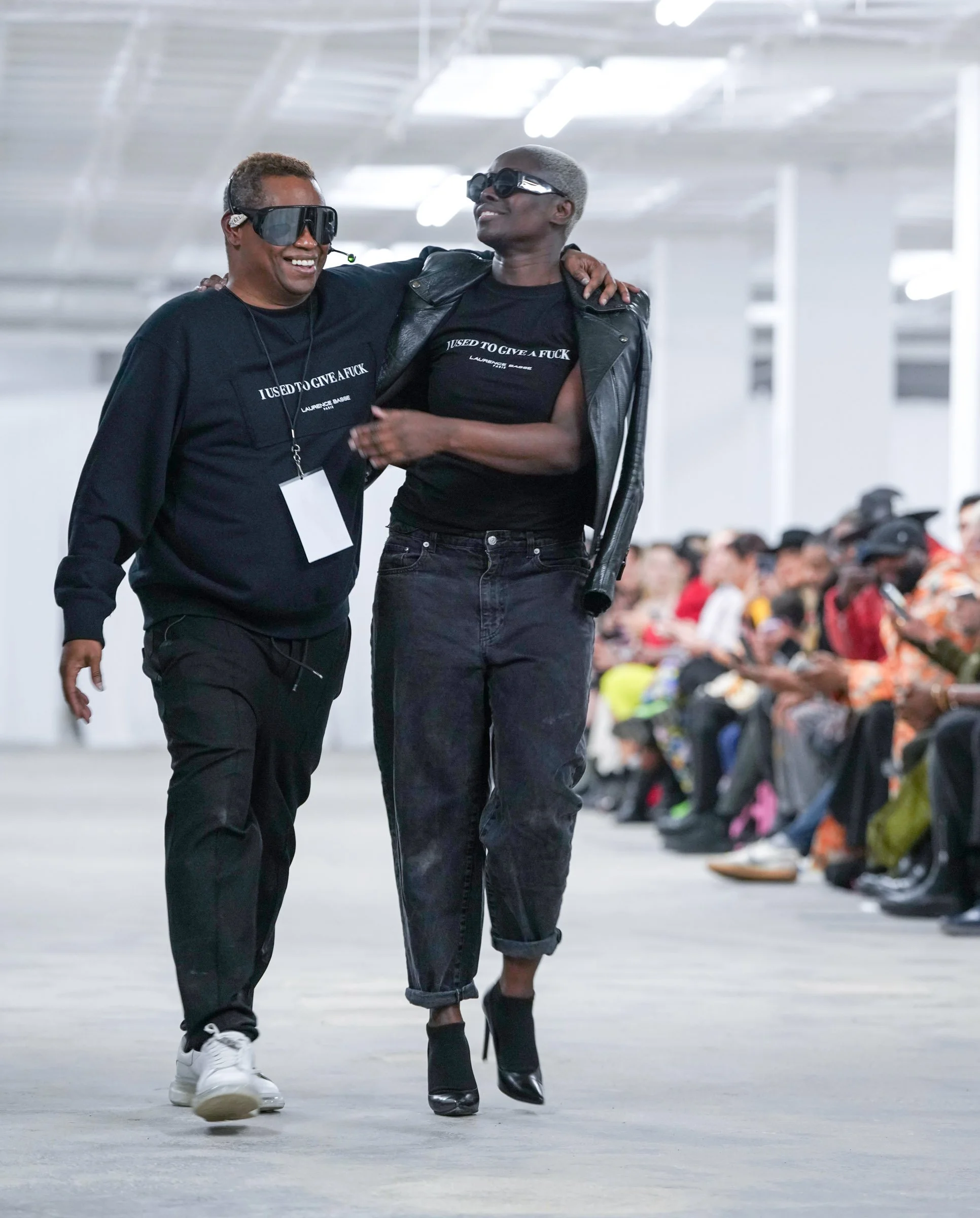 Two people walking together in an indoor space, smiling and wearing sunglasses. The person on the left is dressed in black with a headset, and the person on the right is dressed in black jeans and a leather jacket, with short hair. In the background,