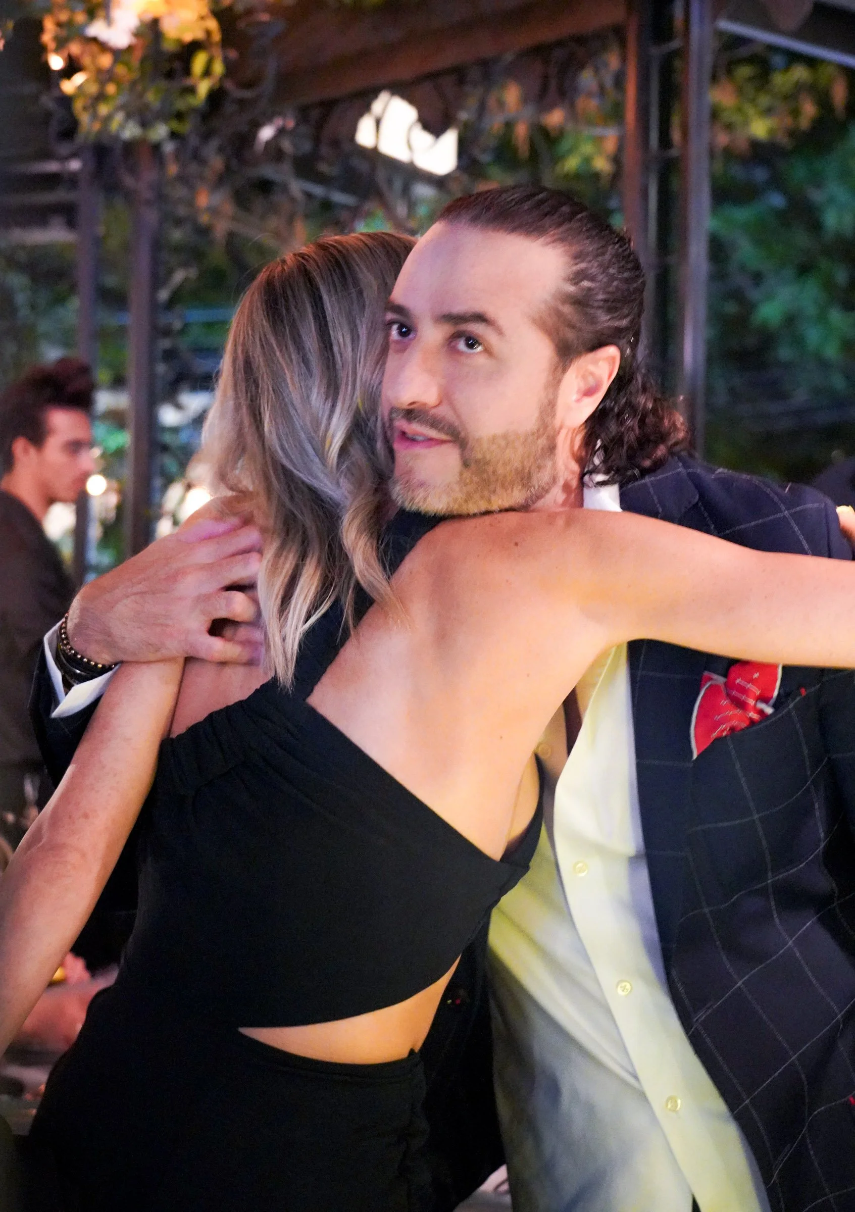 People hugging at a social gathering, with a man in a checkered blazer and a woman in a black dress embracing Isola Ristorante en CDMX.