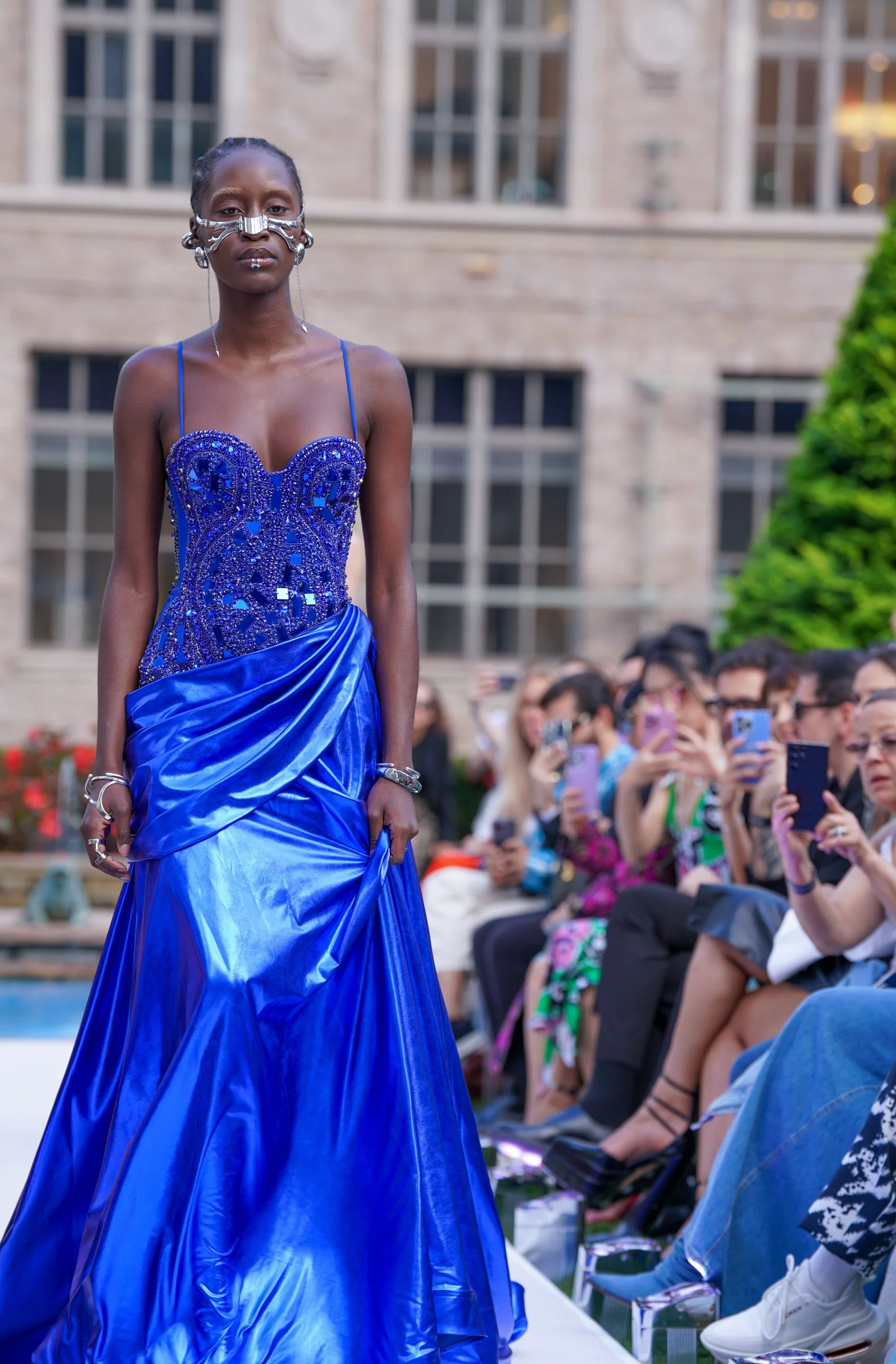 A woman modeled a vibrant blue, embellished gown on an outdoor runway with an audience watching and taking photos.