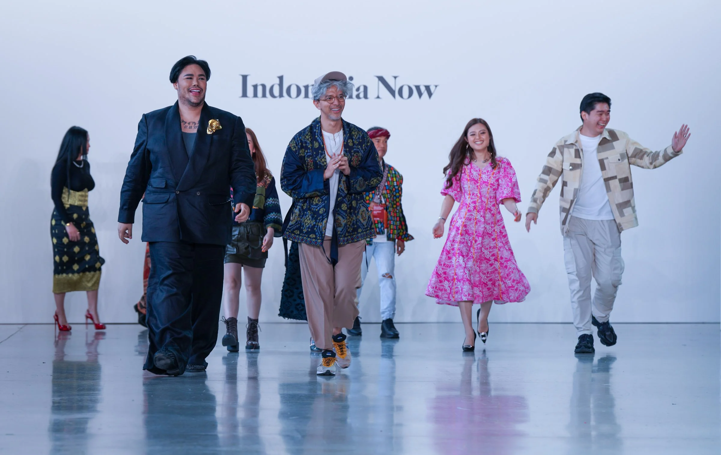 Group of diverse people walking and smiling in a bright indoor space with a white wall in the background that has the text 'Indonesia Now'.