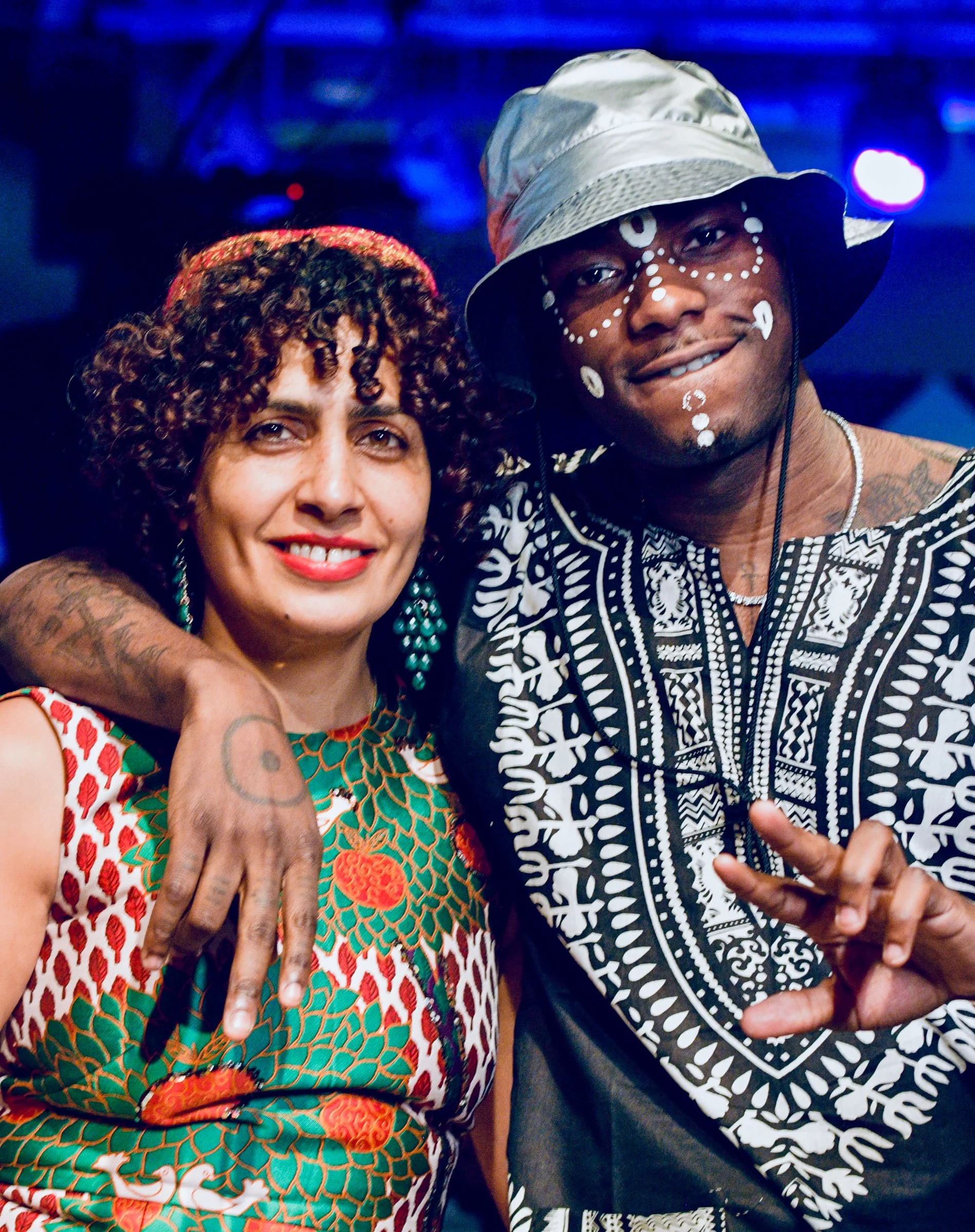 Two people posing together, one woman and one man, at a lively event with stage lighting. The woman has curly hair, is smiling, and is wearing a patterned dress with floral details. The man is wearing a wide-brimmed hat, has face paint or makeup with