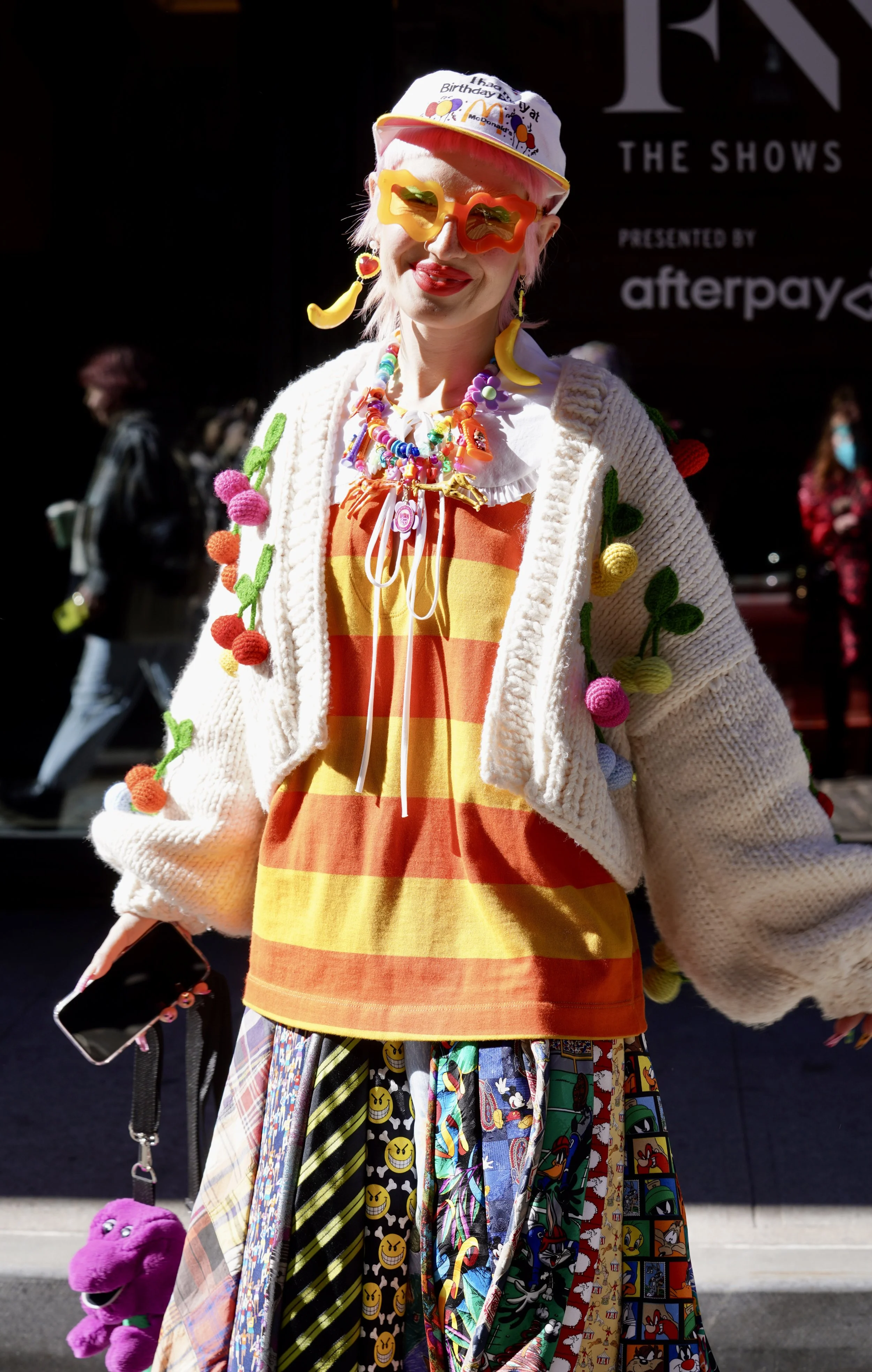 Person dressed in colorful, quirky clothing and accessories, including a white knit cardigan with pom-poms, a striped orange and yellow top, and a skirt covered in cartoon character patches. They wear large, bright yellow sunglasses, a white hat with