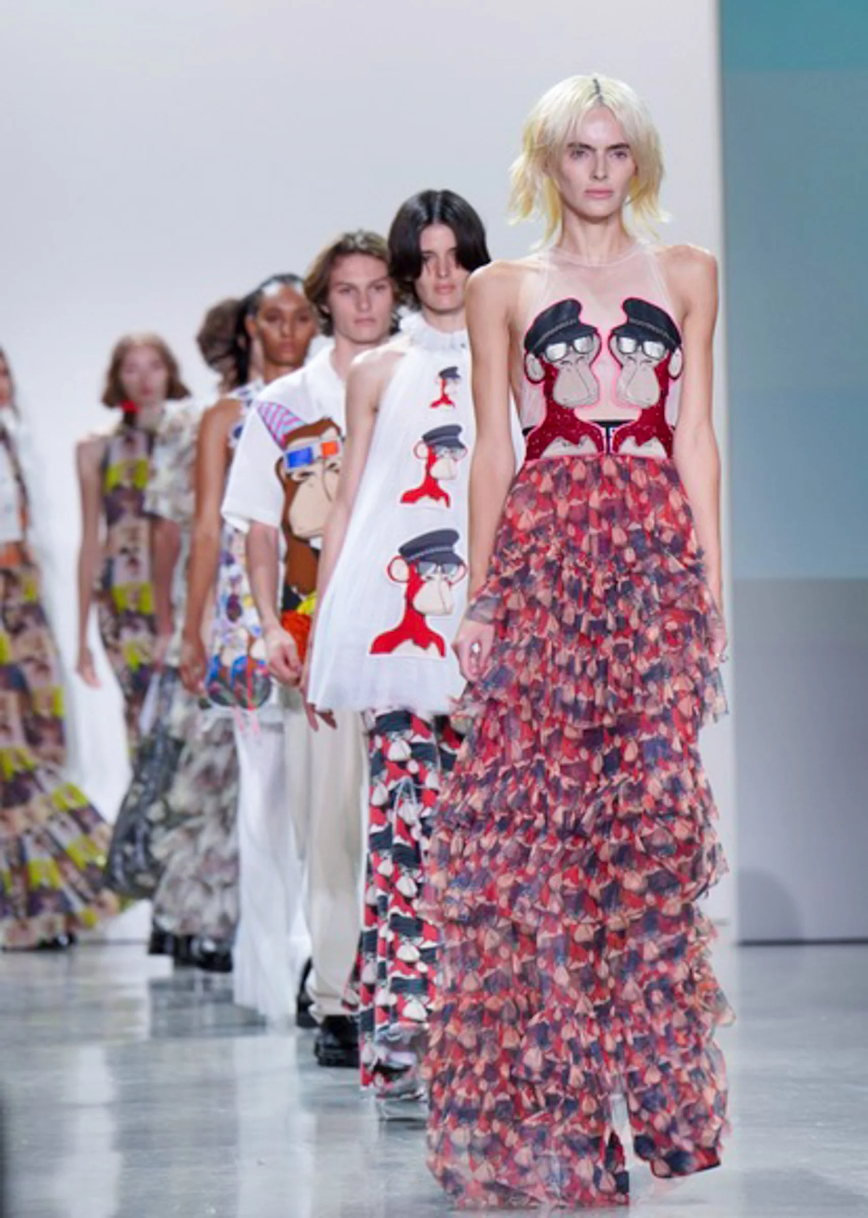 Models walking down a runway during a fashion show, with the leading model wearing a floral patterned dress with a sheer top featuring cartoon characters and the other models wearing various colorful outfits behind her.