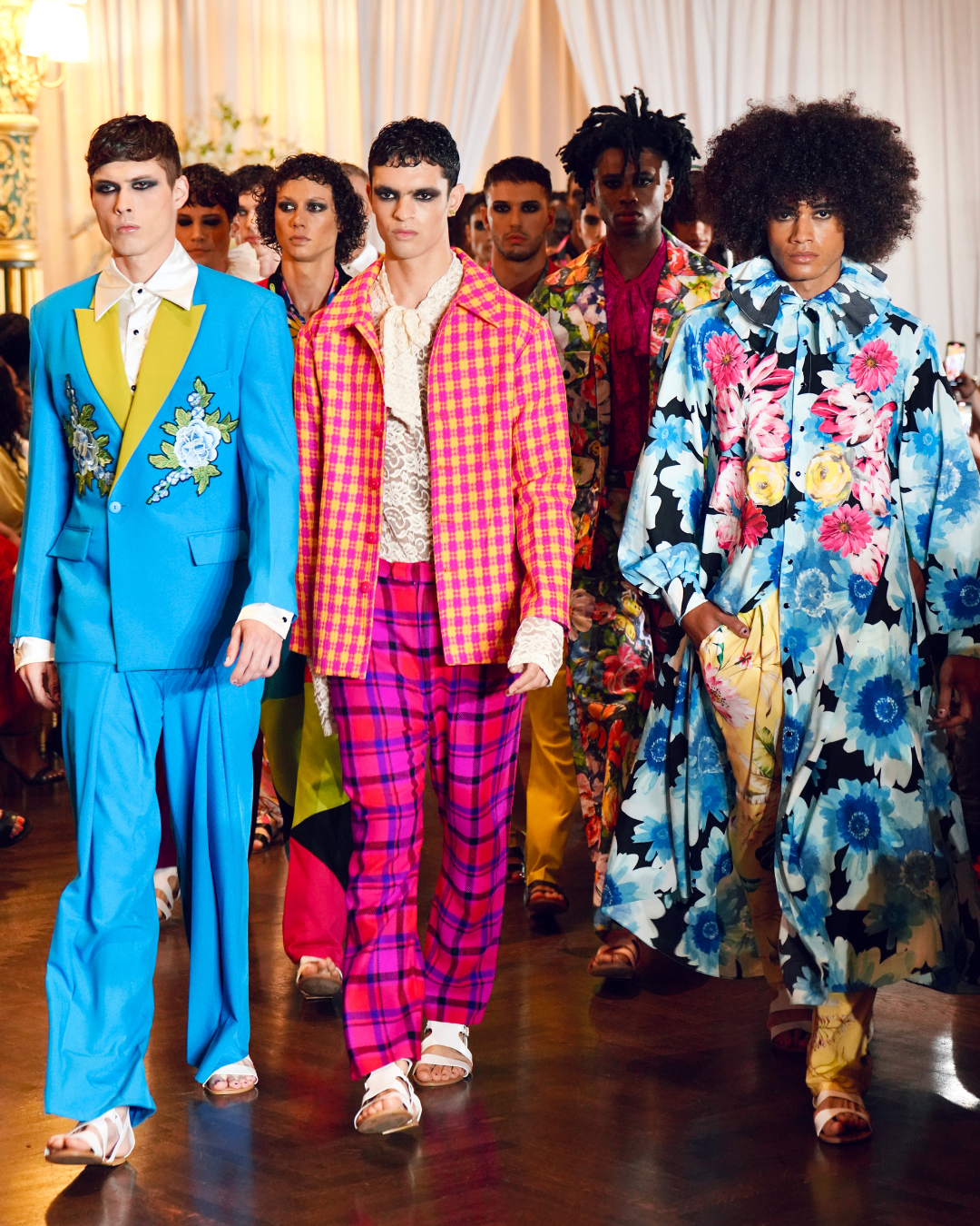Models walking on a runway in colorful floral and patterned suits and dresses with a white tented background.