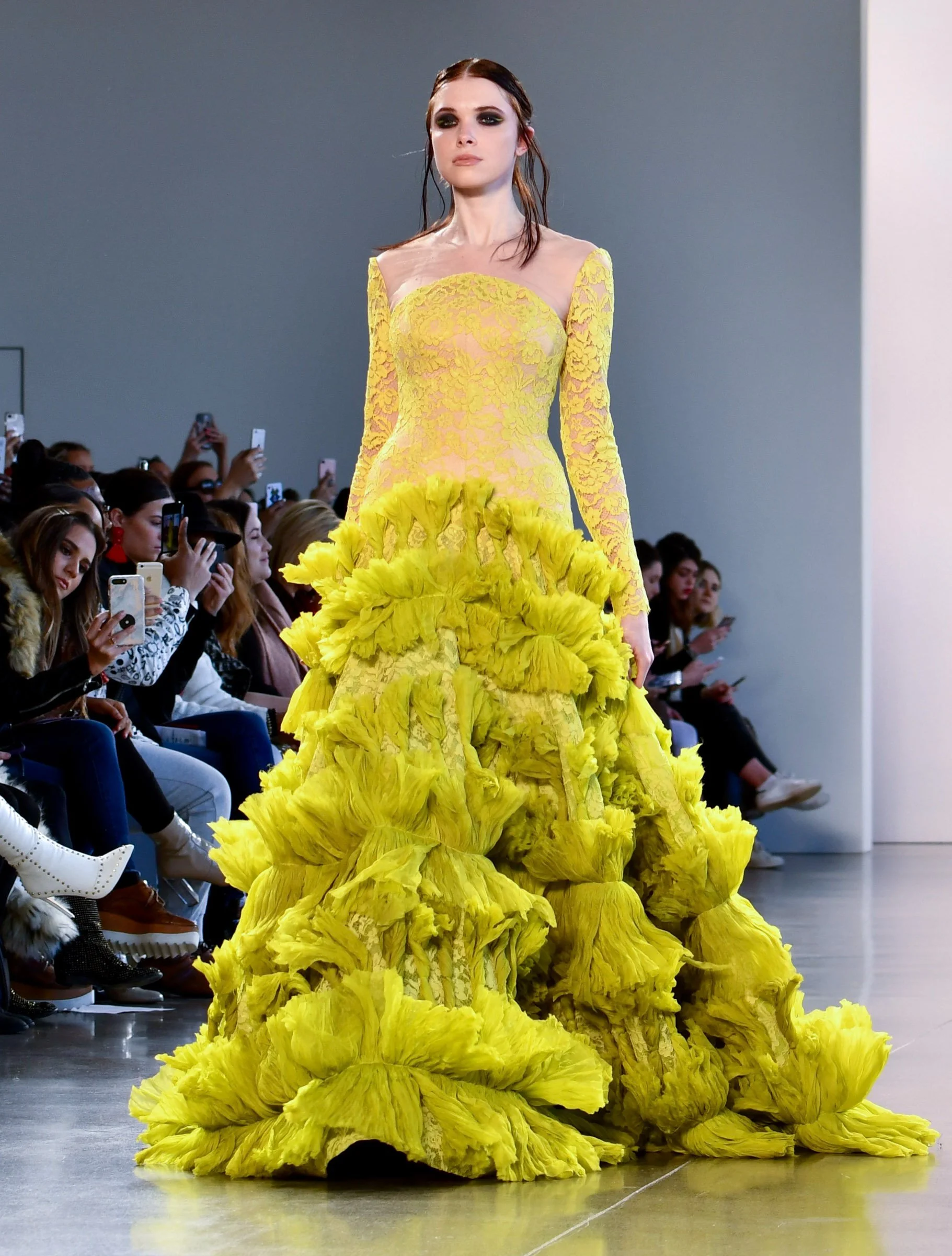 A model walking on a runway wearing a yellow gown with lace details and a full skirt made of ruffled fabric. The audience is seen seated on both sides, many holding up their phones to take pictures.