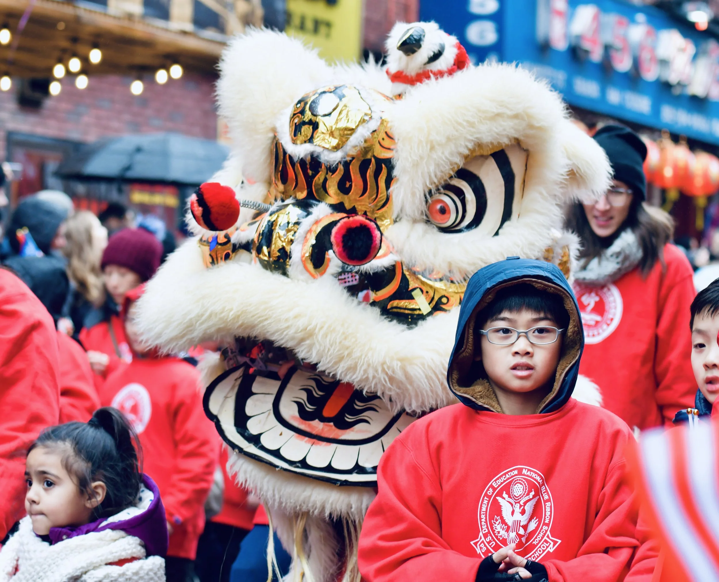 Children participating in a cultural parade in an urban setting with brick buildings and string lights, with a lion dance performer in traditional costume and a lion head, and a blue sign with white text in the background.