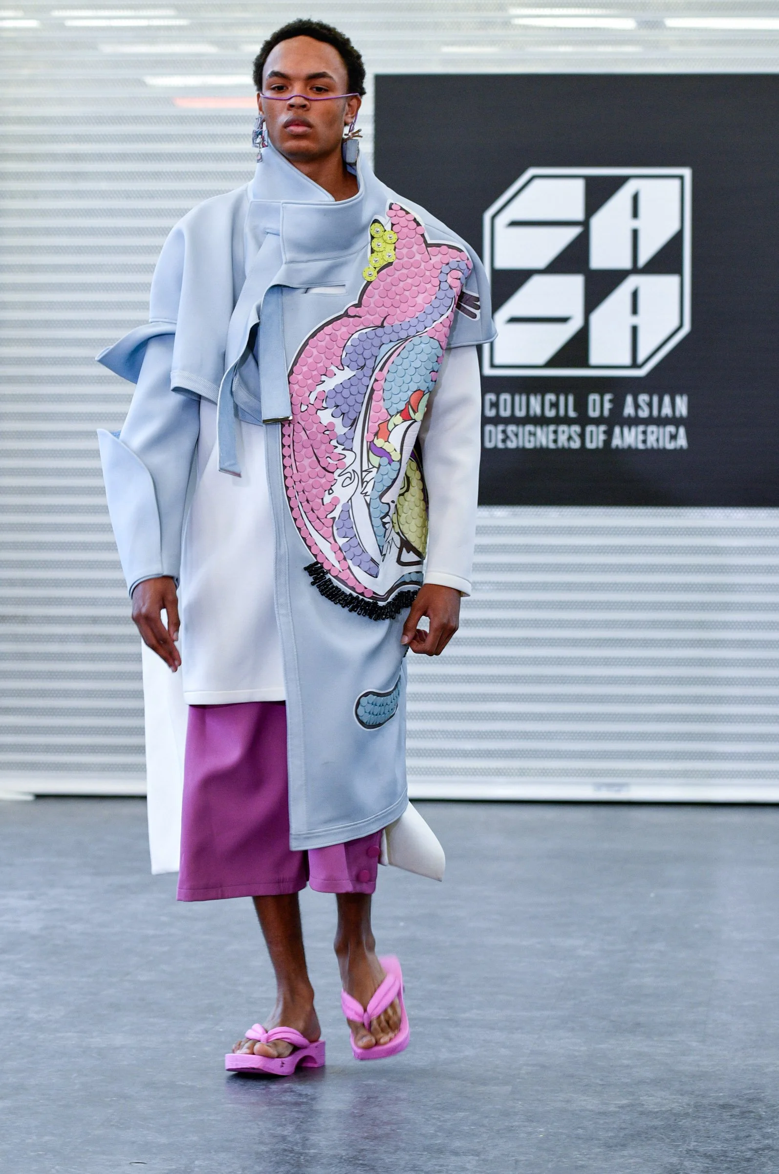 A person walking on a runway wearing a light blue coat with colorful, whimsical embroidery of a pink and purple dragon, paired with purple shorts, pink platform sandals, and pink glasses, at a fashion event for the Council of Asian Designers of Ameri