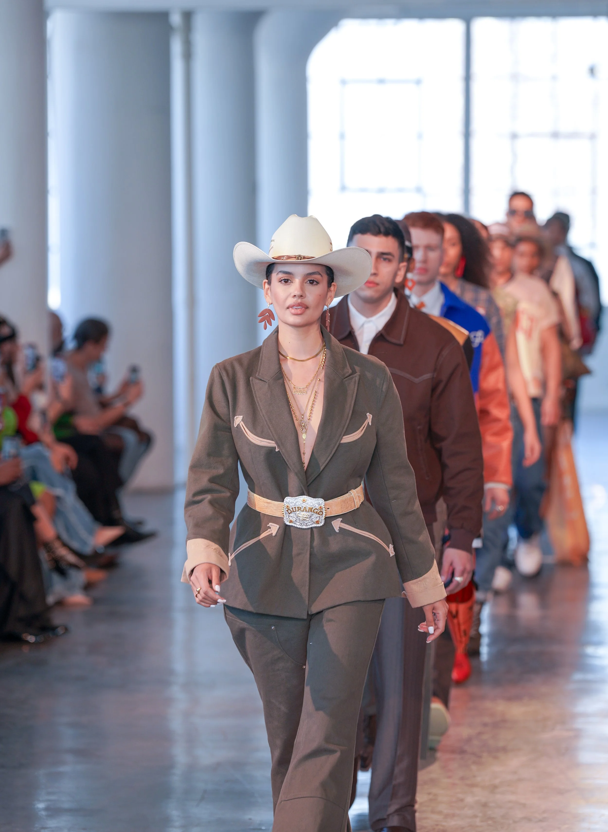 A woman wearing a cowboy hat and western-style clothing walks in front of a line of people on a runway at a fashion show. The background includes large windows and an audience seated along the sides.