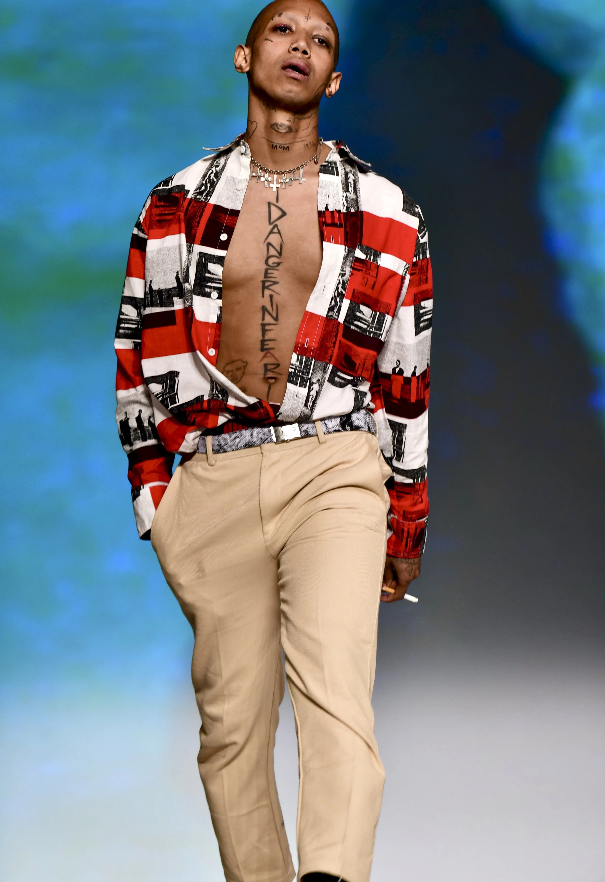 A person with tattoos and pierced ears walks on a runway, wearing beige pants and a partially open red, black, and white patterned shirt, against a blue and green gradient background.
