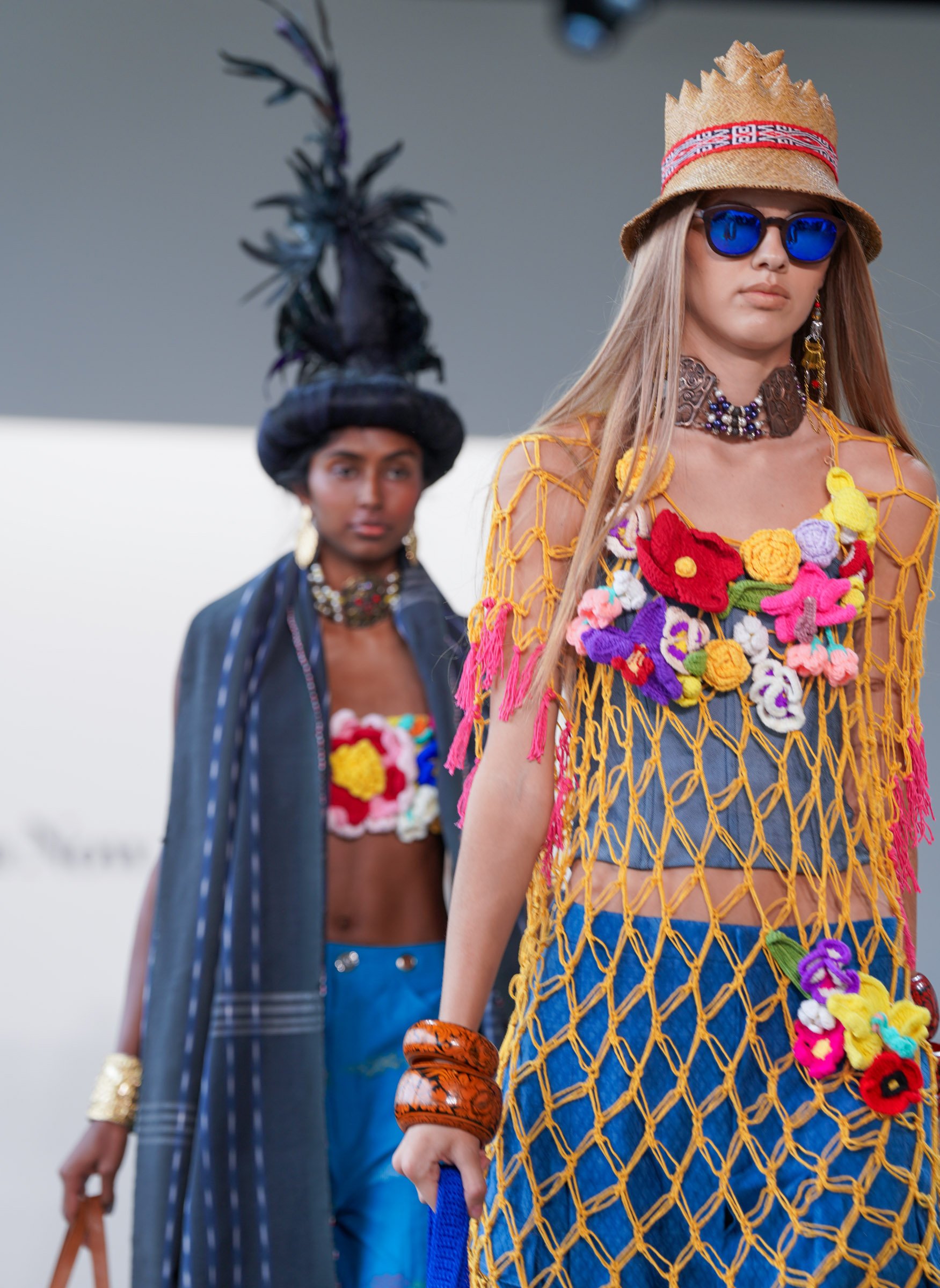 Fashion models walking on a runway wearing colorful, eclectic outfits with accessories, including hats, sunglasses, and jewelry.