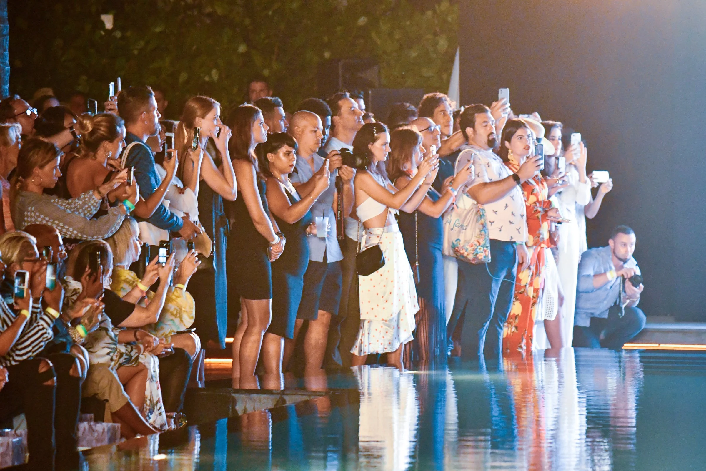 A diverse group of people standing closely together, watching a performance or event at night, with some taking photos or videos with their smartphones.