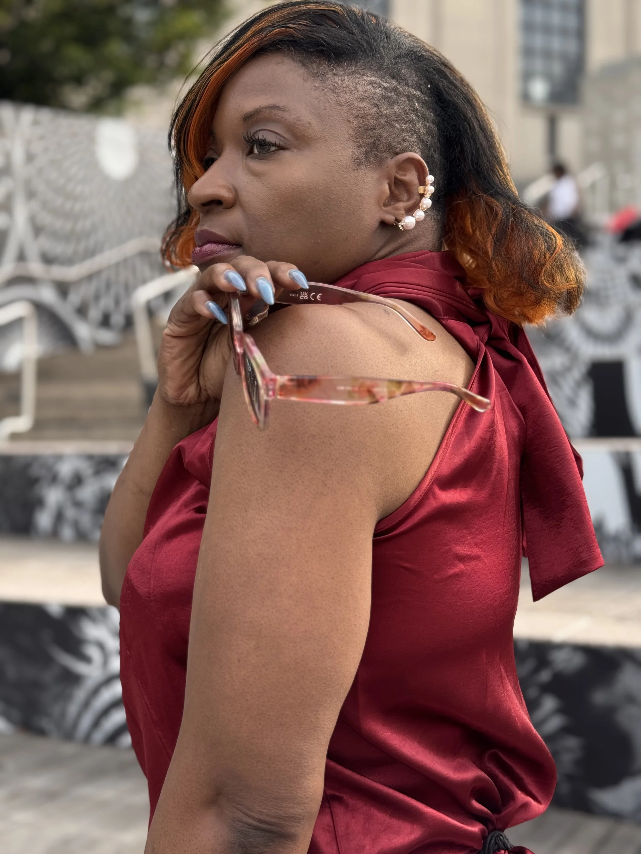 A woman with short, asymmetrical hair styled with orange highlights and partial shaved sides, wearing a red satin dress, pearl earrings, and holding glasses, posing outdoors in an urban setting.
