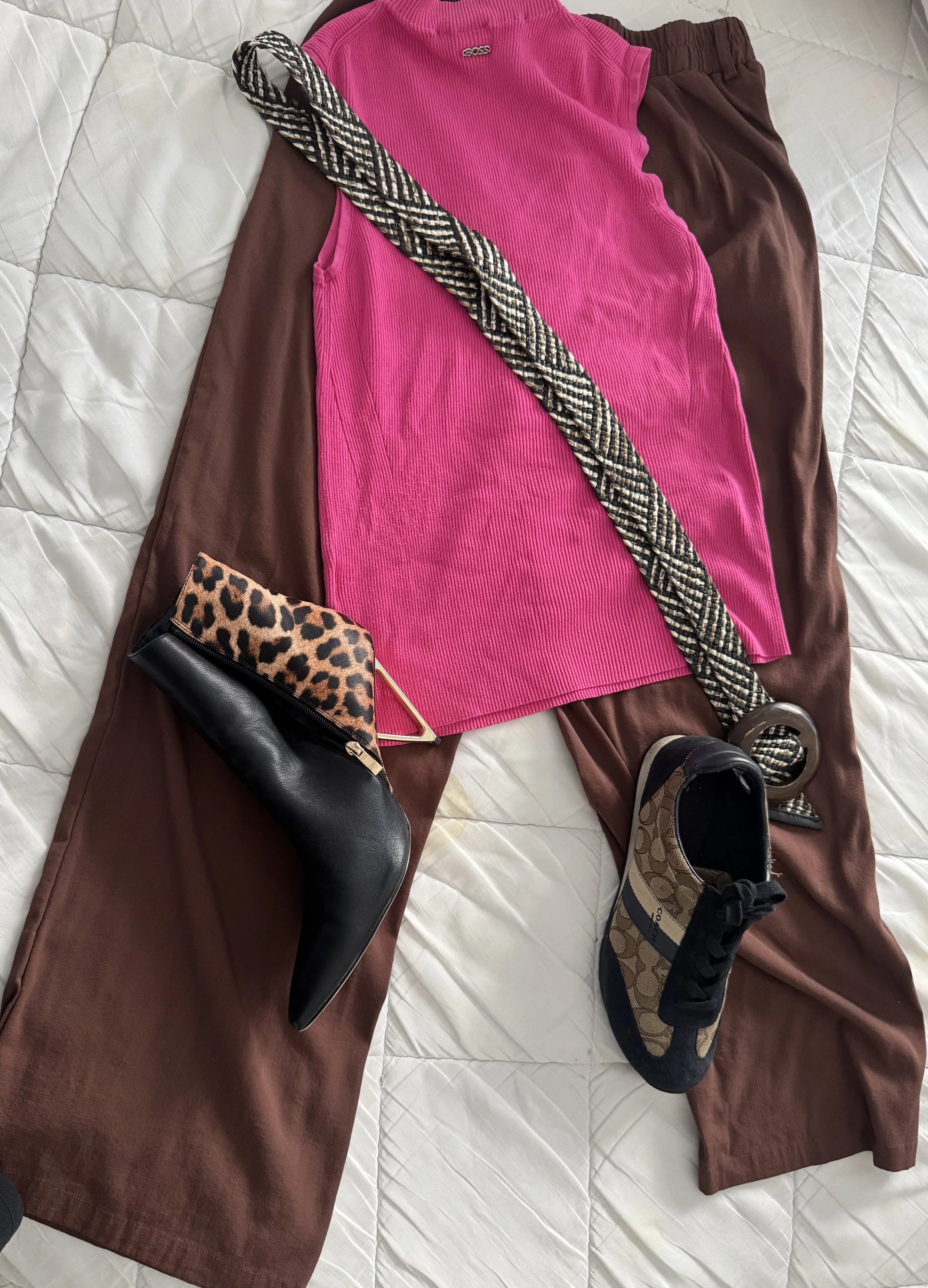Clothing and accessories laid out on a bed, including brown pants, a bright pink ribbed sleeveless top, black and leopard print purse, a woven belt, and a pair of Coach sneakers.