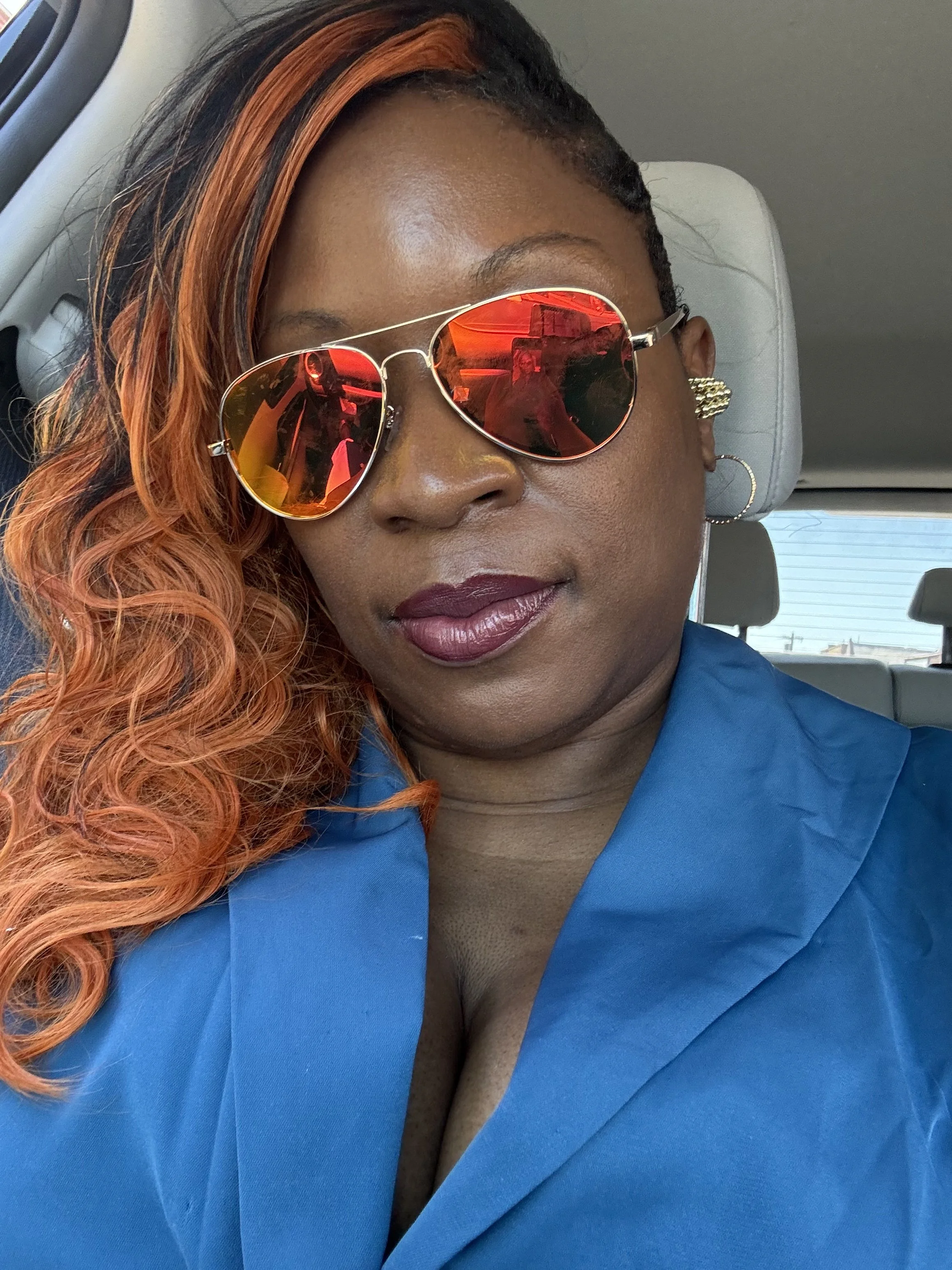 Close-up photo of a woman with braided hair, wearing mirrored sunglasses and a blue blazer, sitting in a vehicle.