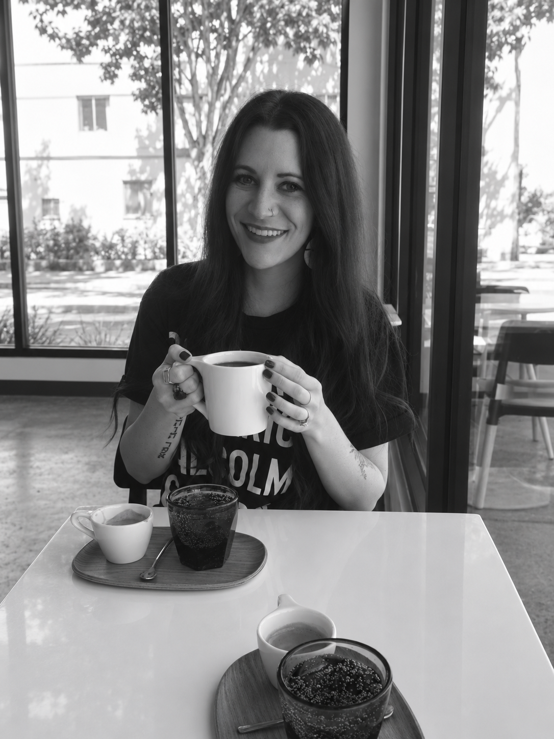 Black and white photo of a woman with long dark hair, smiling, holding a white mug of coffee or tea, sitting at a table in a cafe with large windows and trees outside.
