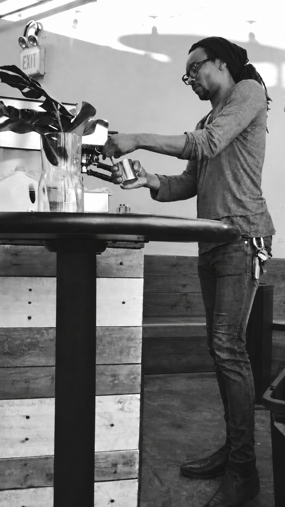 A man with glasses, a headscarf, and a long-sleeve shirt is making coffee with a manual espresso machine in a coffee shop, with a large plant in a glass vase on the counter.
