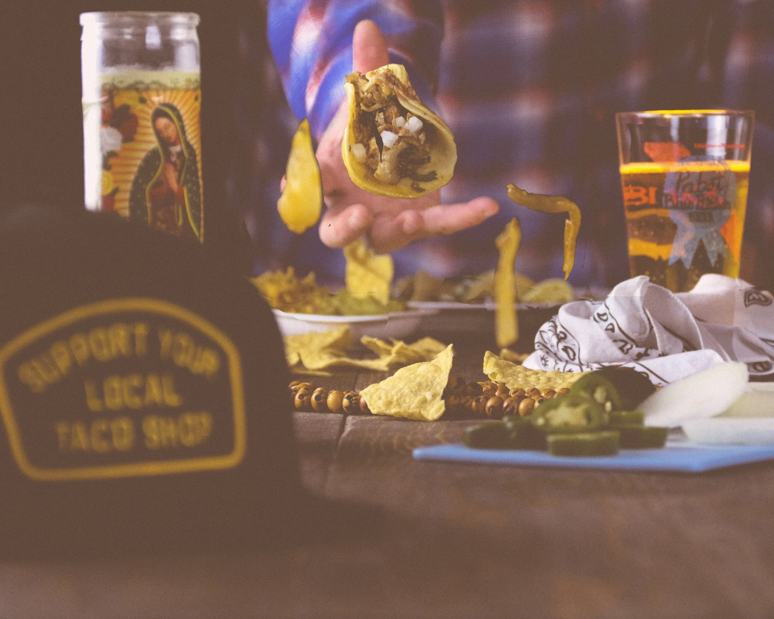 A hand holding a taco with chopped meat and diced onions, surrounded by taco shells, a beer glass, and various taco toppings on a table.