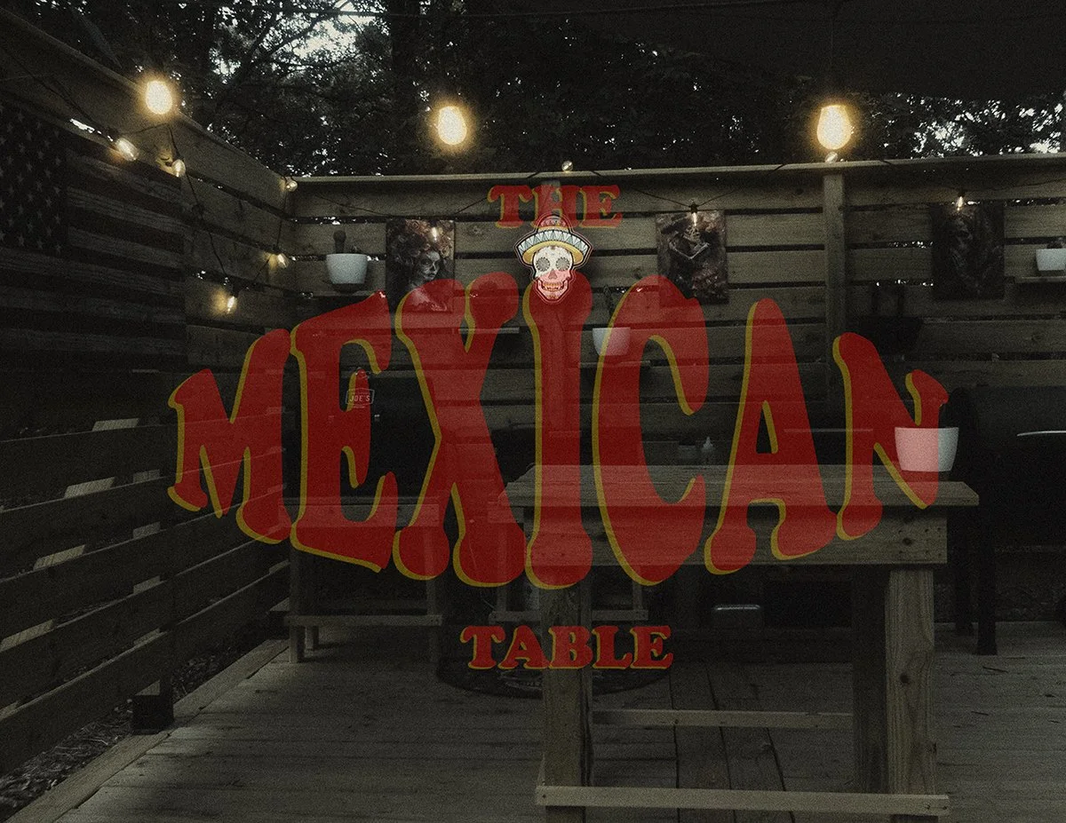 A wooden sign with large red and yellow letters spelling "The Mexican Table" hangs on a wooden fence at an outdoor setting with string lights, potted plants, and a dark sky in the background.
